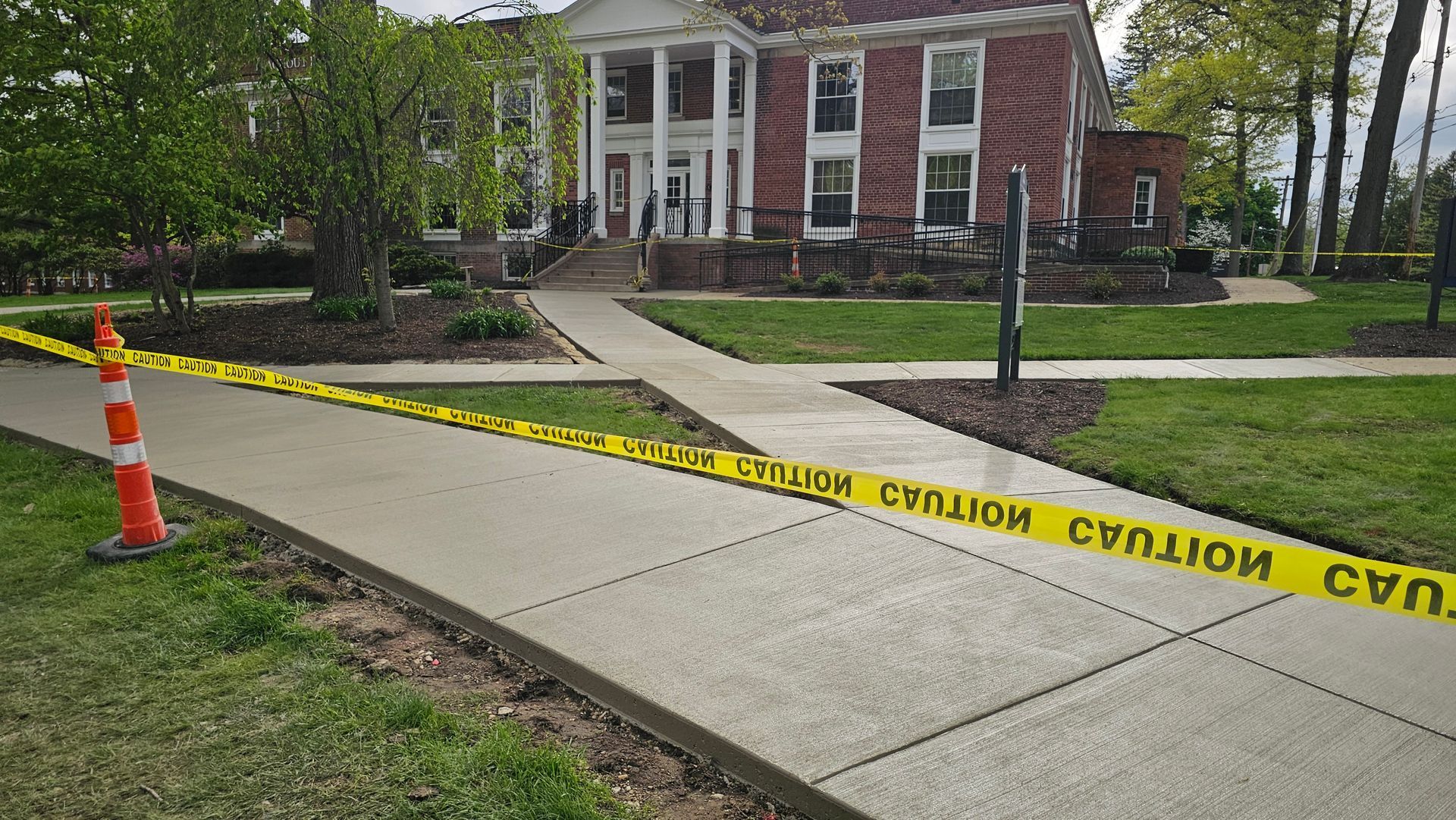 A brick building with white columns behind a sidewalk cordoned off with yellow caution tape and an orange traffic cone.