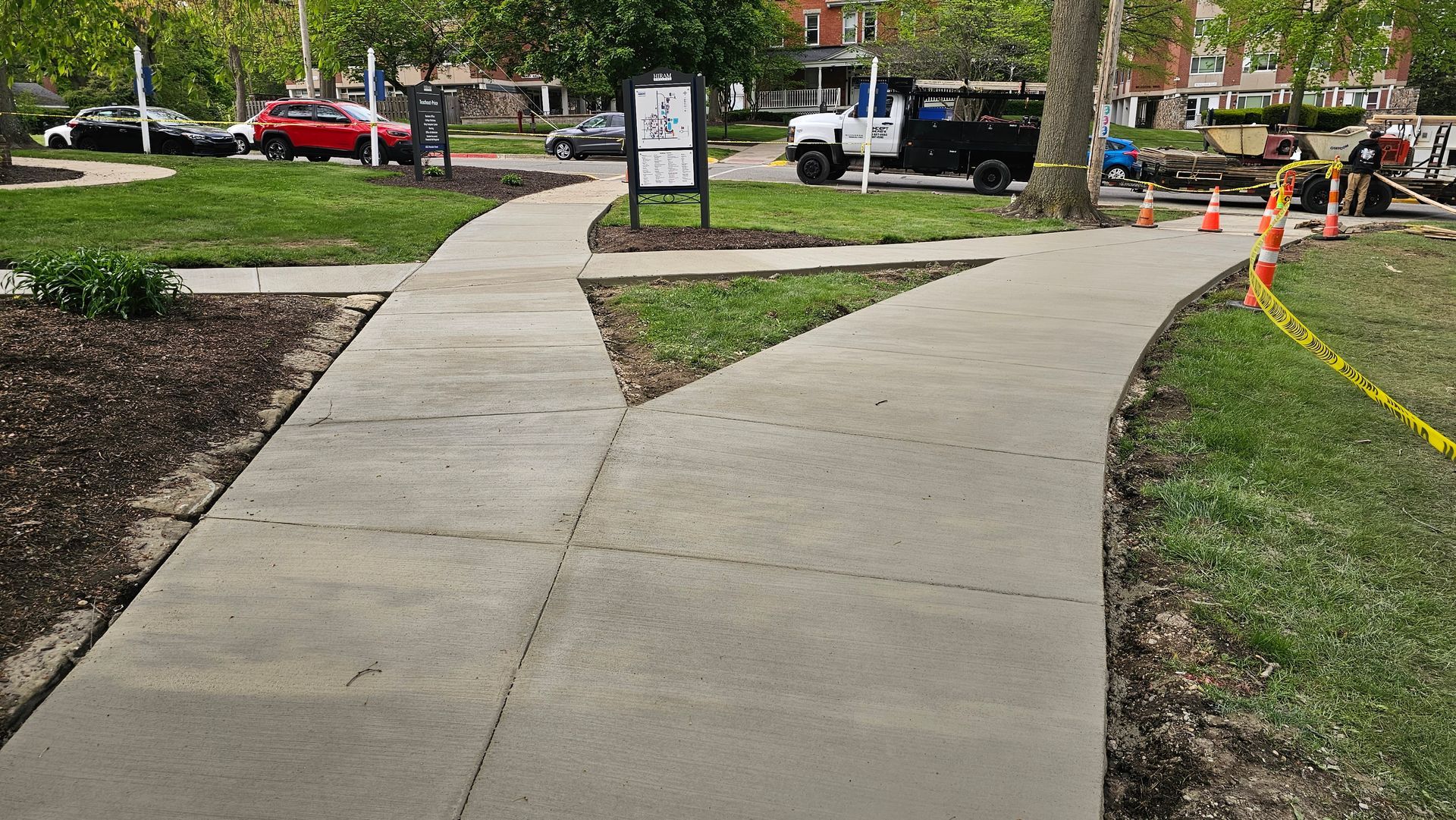 A concrete walkway forks into two paths in a park, with orange construction cones and caution tape nearby.
