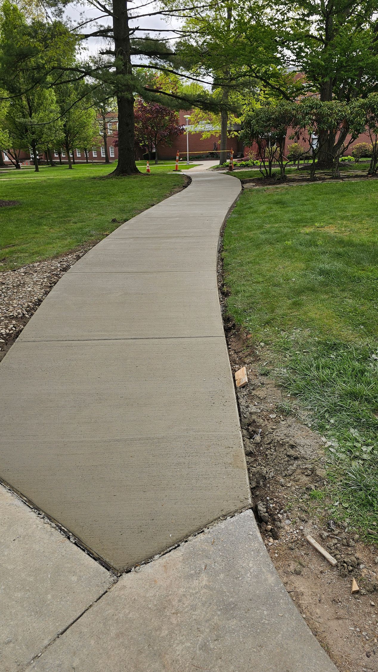 A curved concrete sidewalk leads through a park toward a brick building surrounded by trees and green grass.