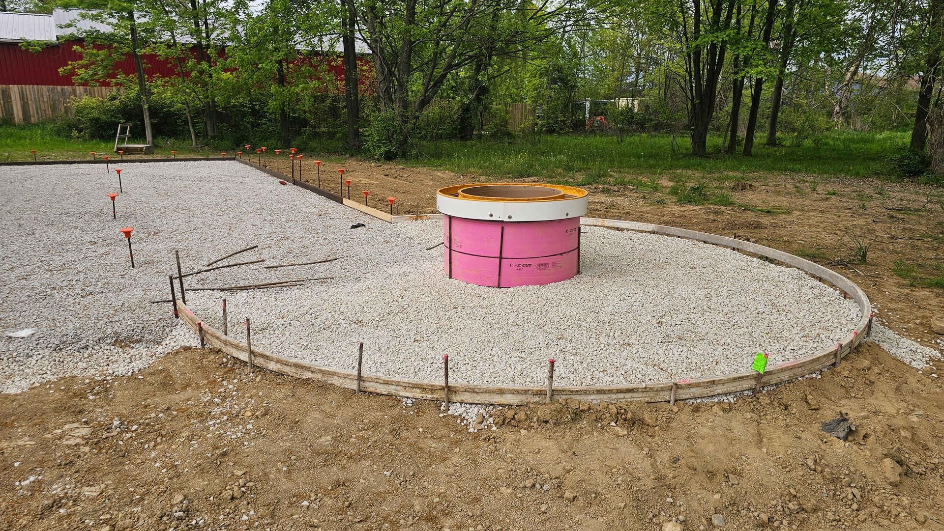 A construction site shows a gravel area with wood forms and a pink cylindrical foam mold for a concrete foundation.