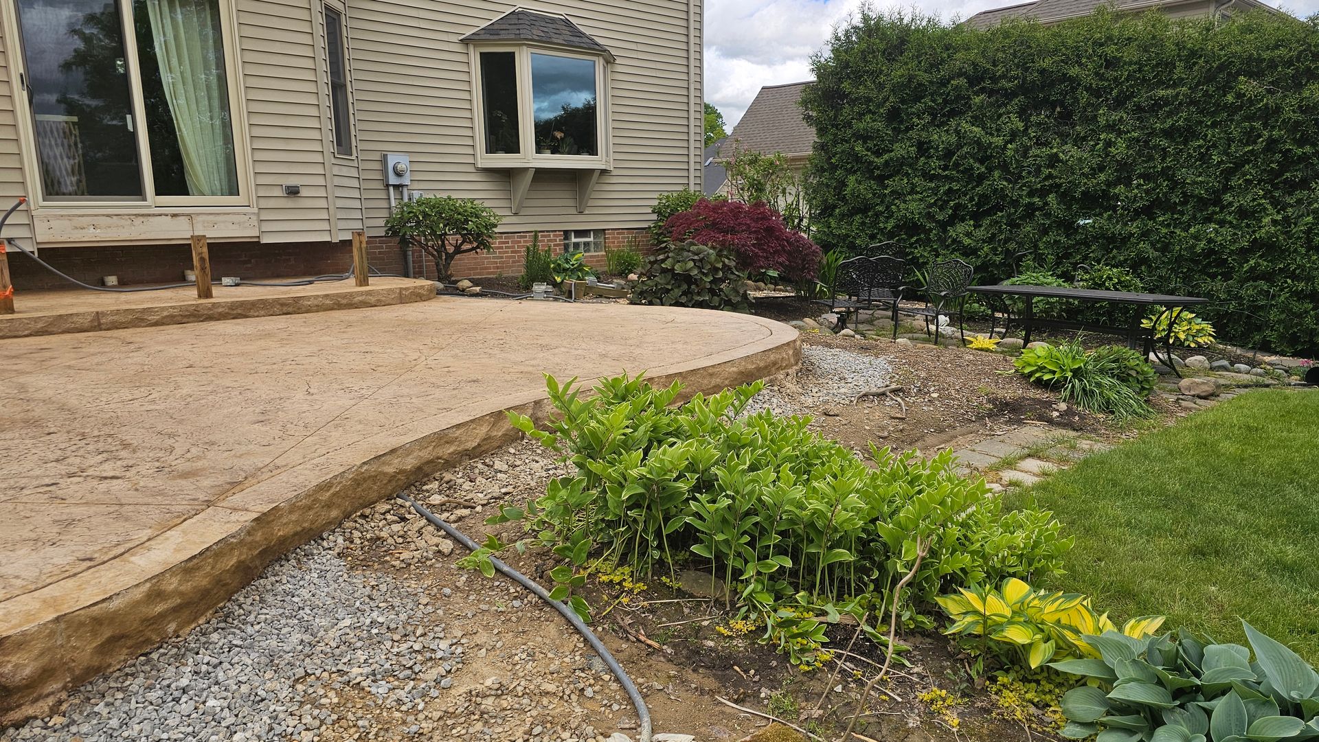 A tan stone patio sits beside a house, next to a landscaped garden bed with green plants, small rocks, and a hedge.