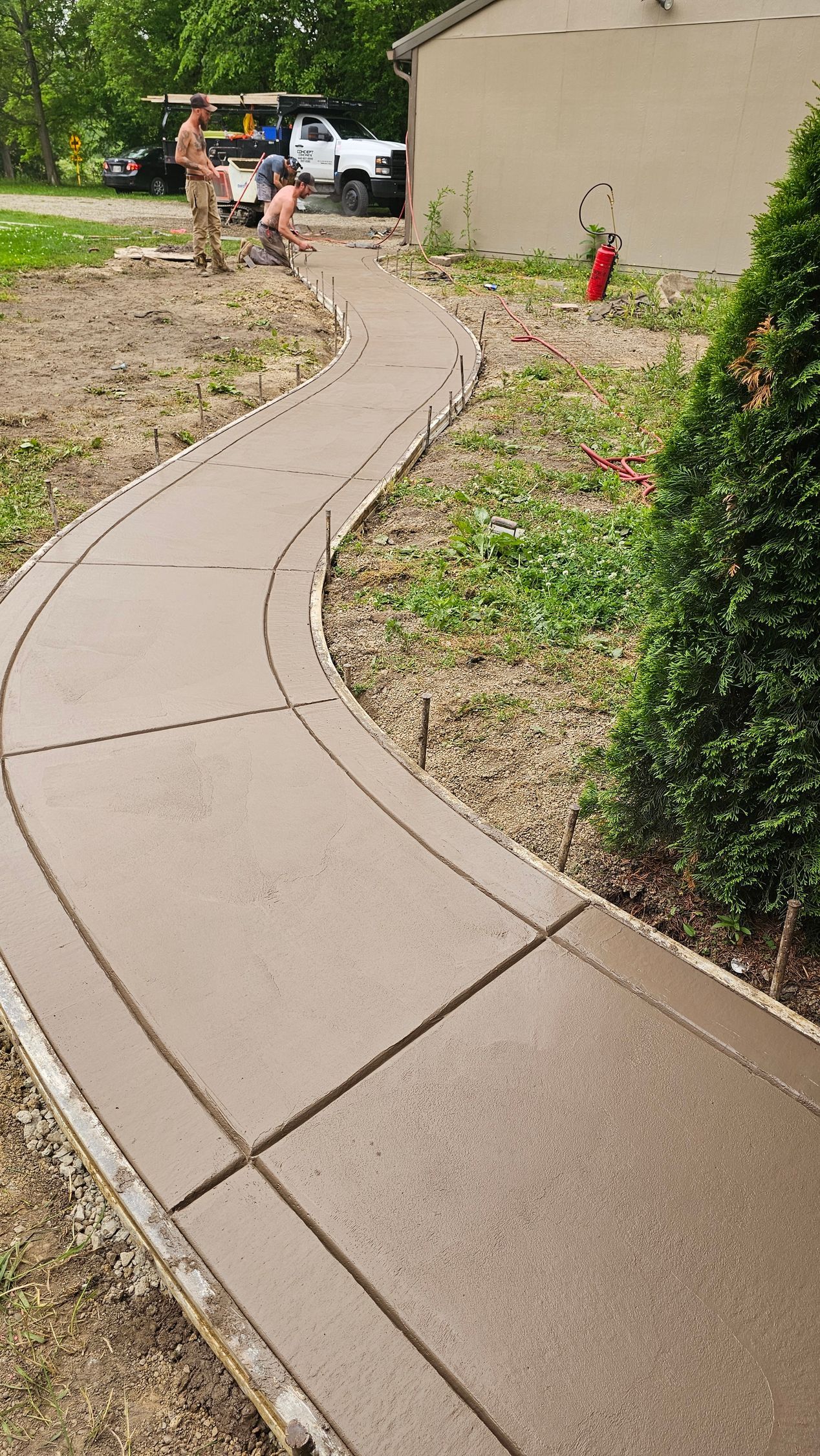 A newly poured concrete pathway curves through a yard next to a building, with two workers in the background.