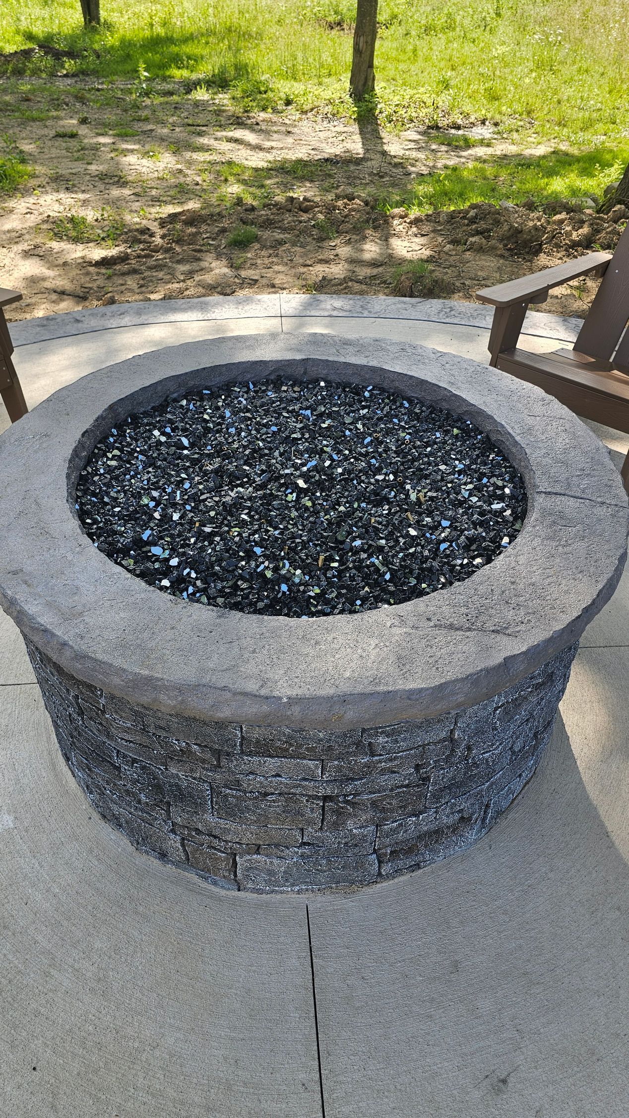 A circular concrete fire pit filled with dark glass pebbles on a stone patio outdoors.