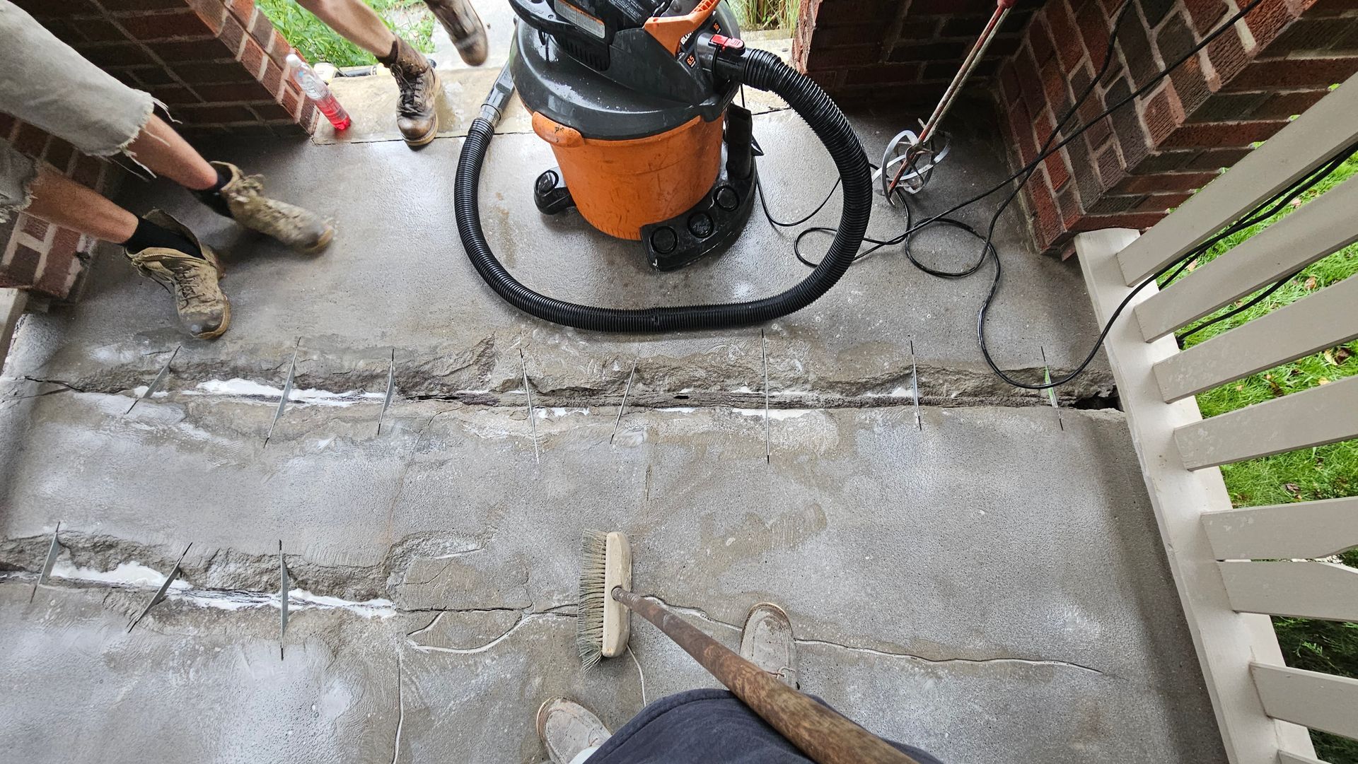 A person holds a long-handled brush on a concrete porch, cleaning dust from deep cracks alongside a shop vacuum.