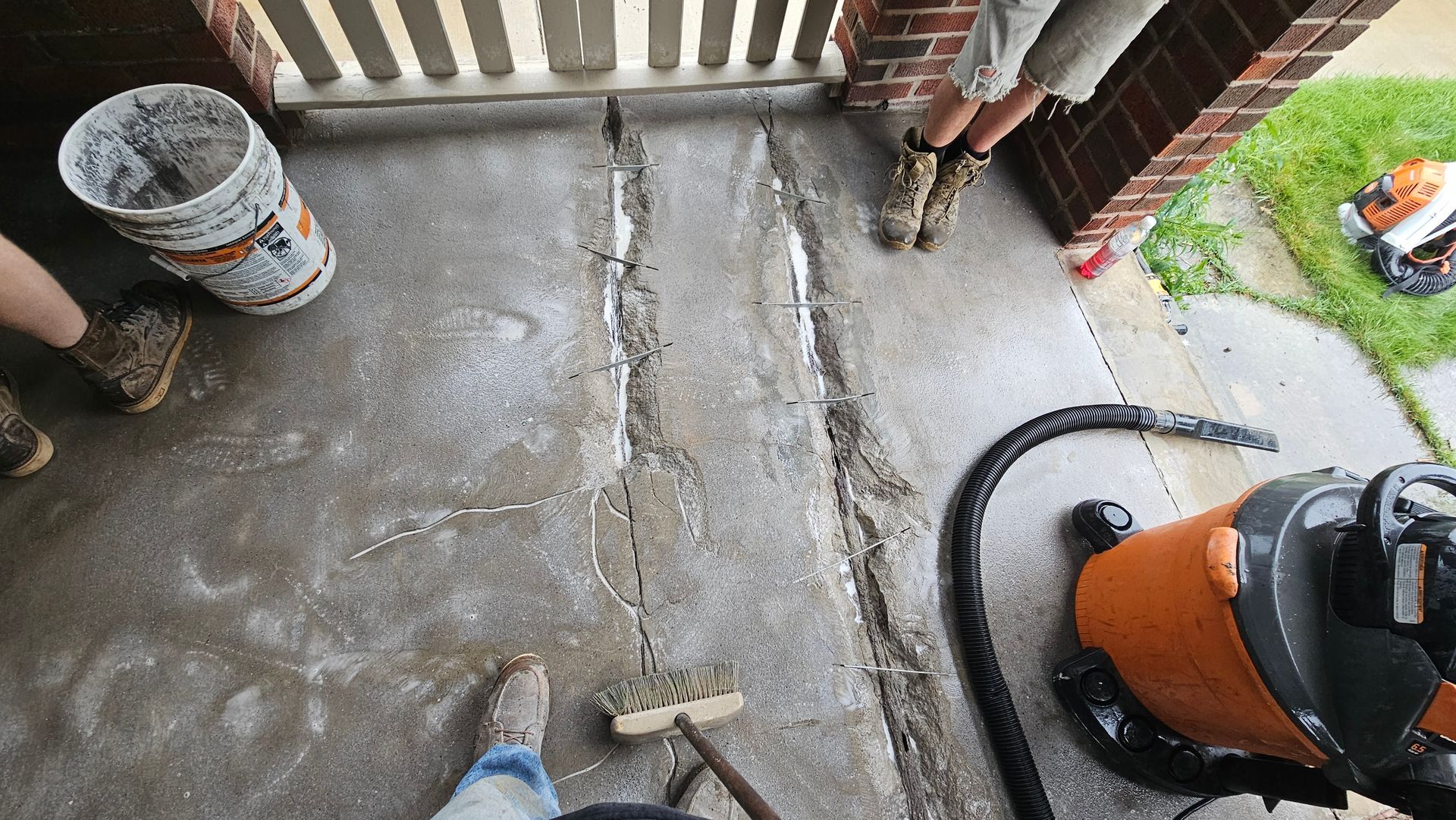 A person prepares to repair a long, vertical crack in a concrete porch, with a bucket, vacuum, and tools nearby.