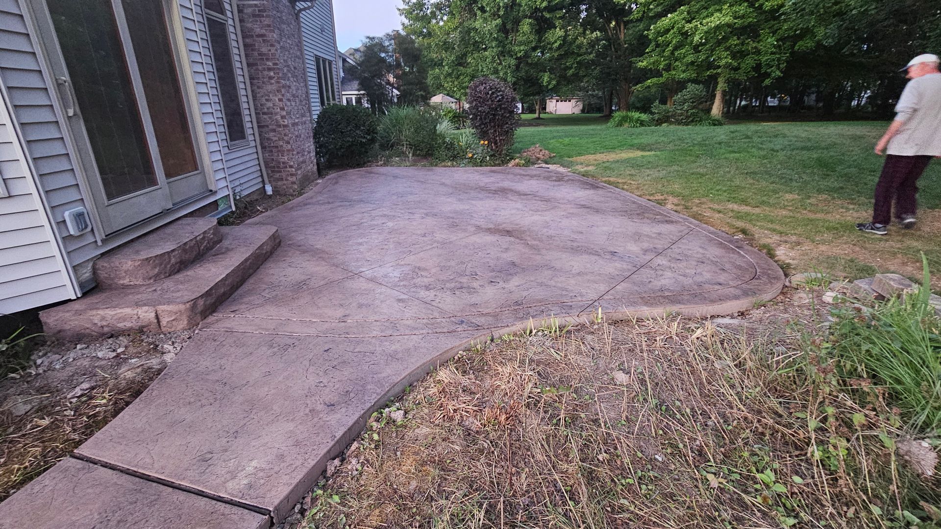 An outdoor stamped concrete patio and walkway adjacent to a residential home with a person standing on the grass nearby.