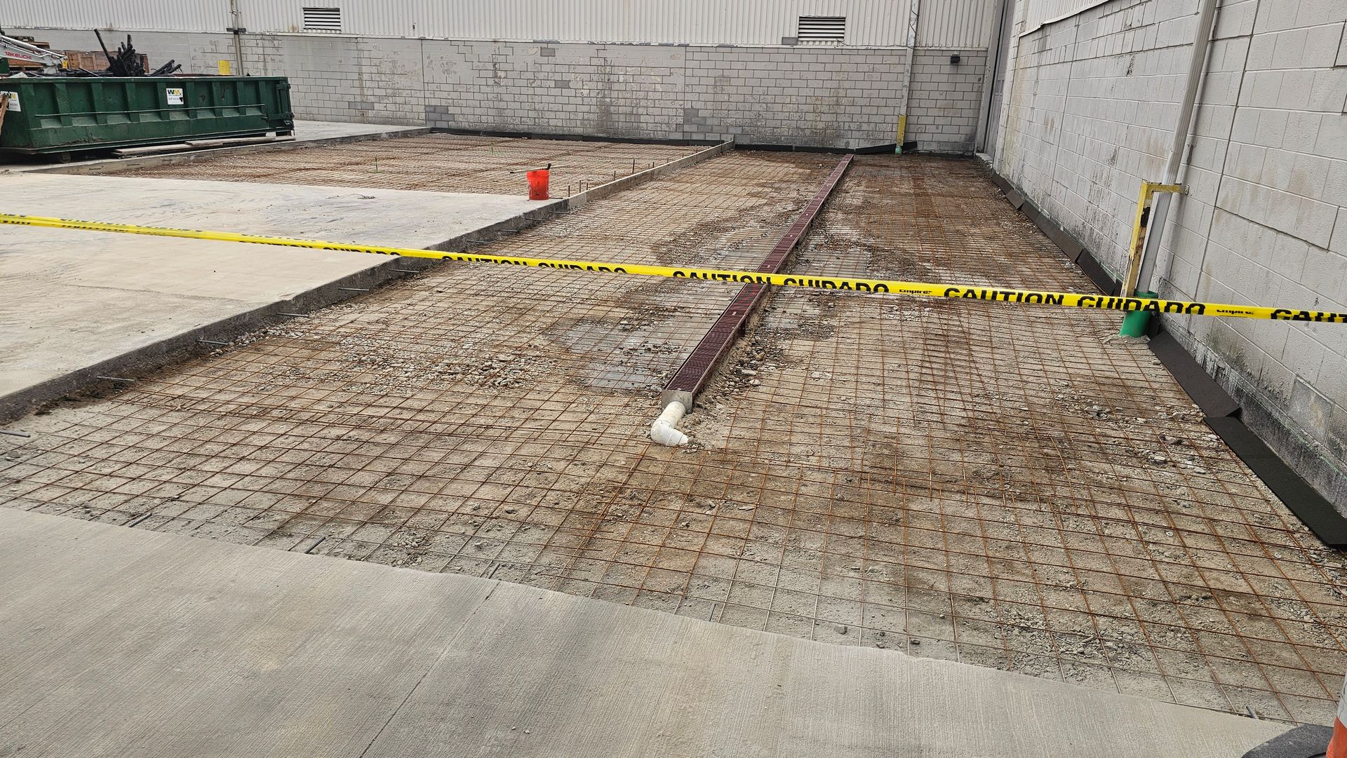 A construction site showing a dirt-filled area prepared for concrete, featuring wire mesh, a red pipe, and caution tape.