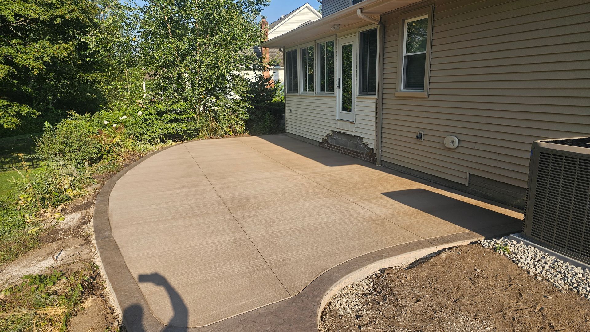 A concrete patio with a dark, curved border sits beside the tan siding of a house, leading to a door and a yard.