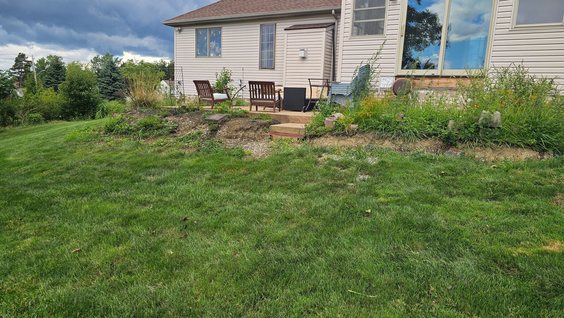 A light-colored house with a patio area and lawn, framed by lush green plants under a cloudy, dramatic sky.