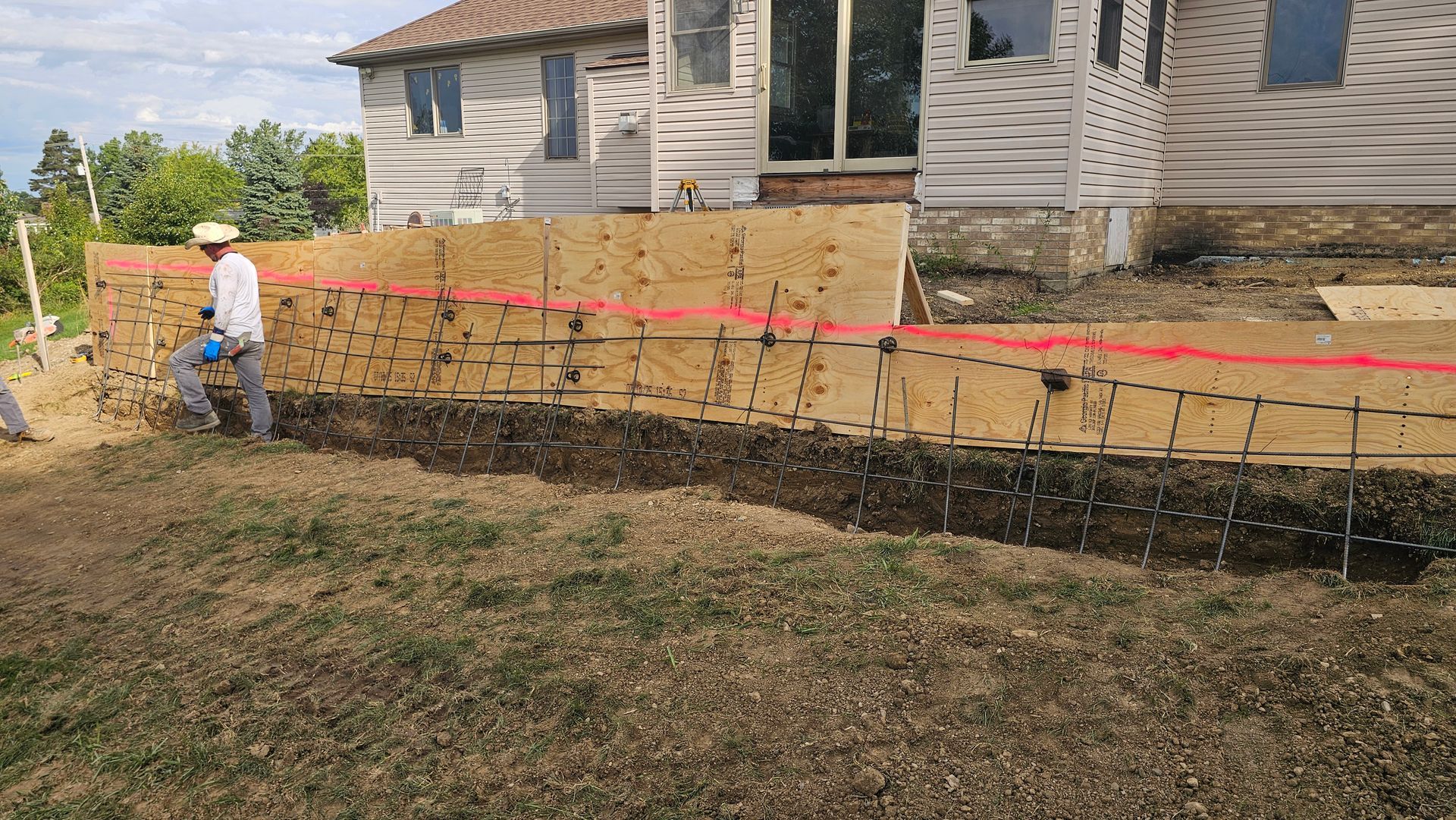 A person works near wooden concrete forms and steel rebar for a foundation wall outside a house.