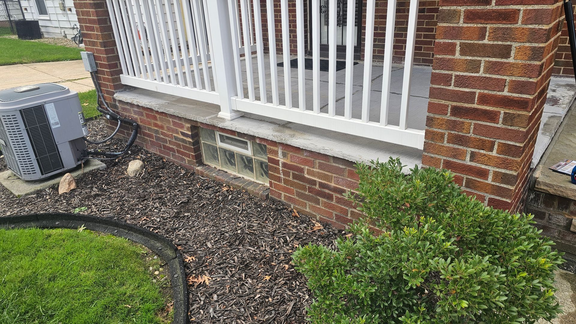 A red brick porch base with white railing, a glass block window, and an air conditioning unit in a mulched garden.
