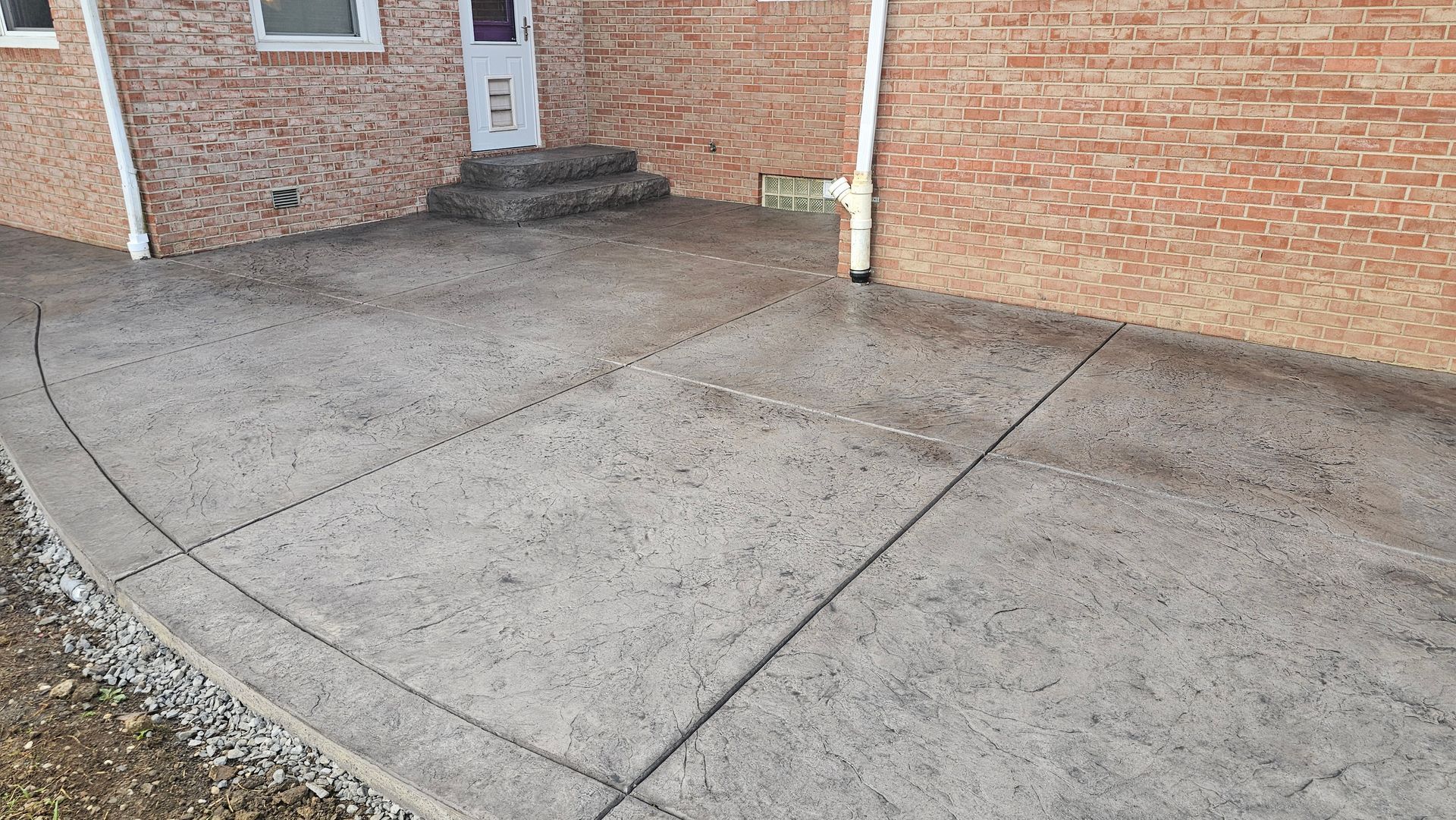 A light-colored brick building with a concrete patio featuring visible control joints and stairs leading to a door.