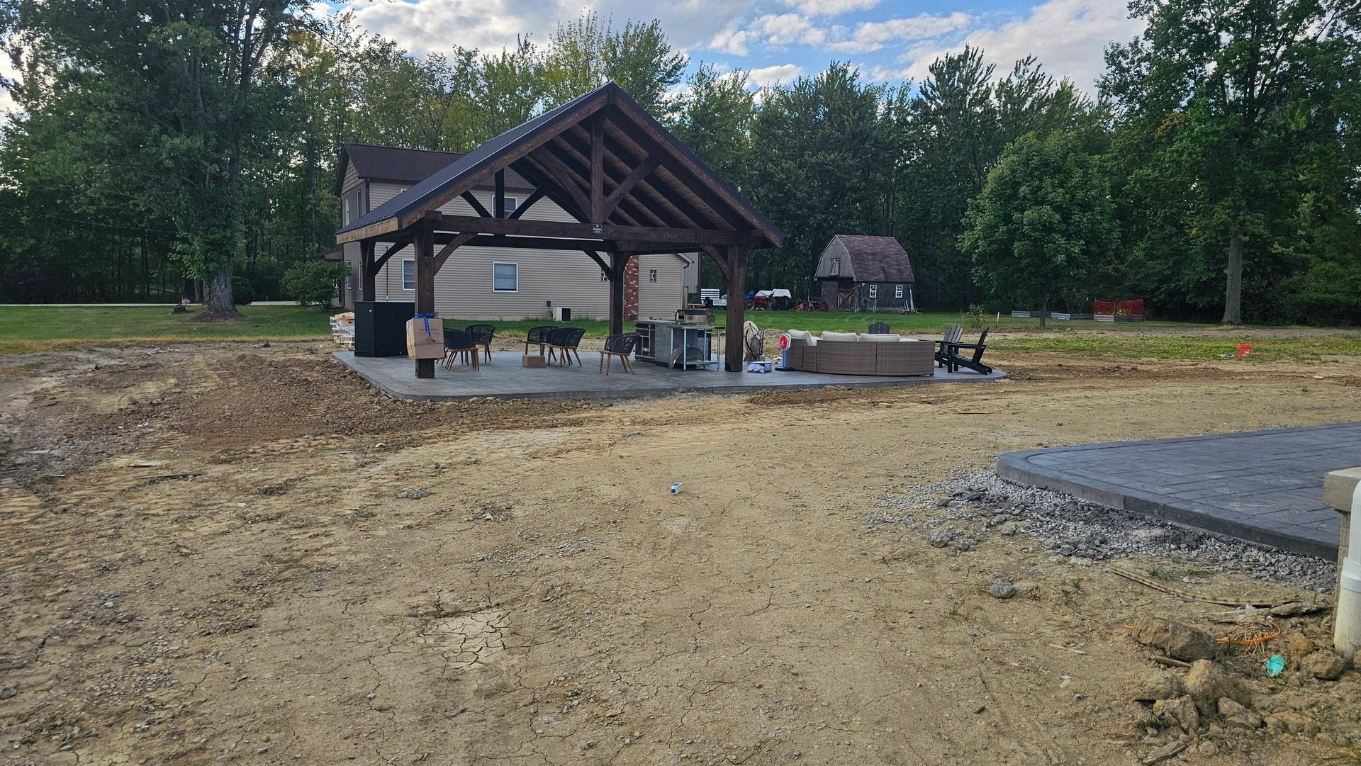 A wooden pavilion with a dark shingled roof stands on a concrete patio in a backyard under a blue, cloudy sky.