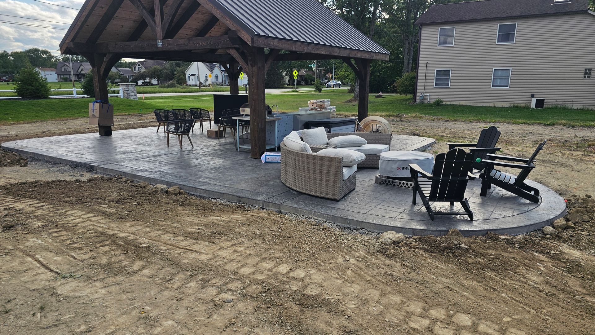 A covered outdoor patio with a stone-textured floor, featuring a curved sofa, fire pit, and chairs in a yard under construction.