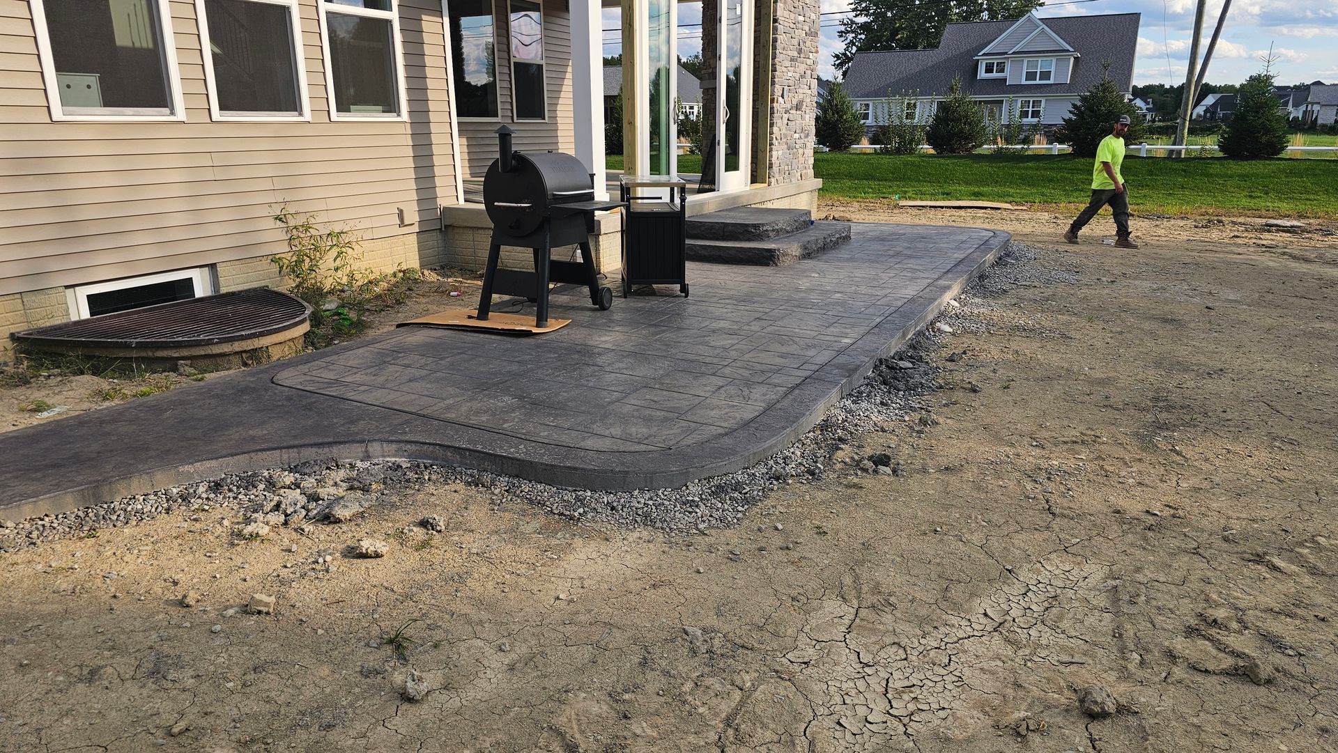 A freshly poured stamped concrete patio outside a house, with a grill and a person walking in the background.