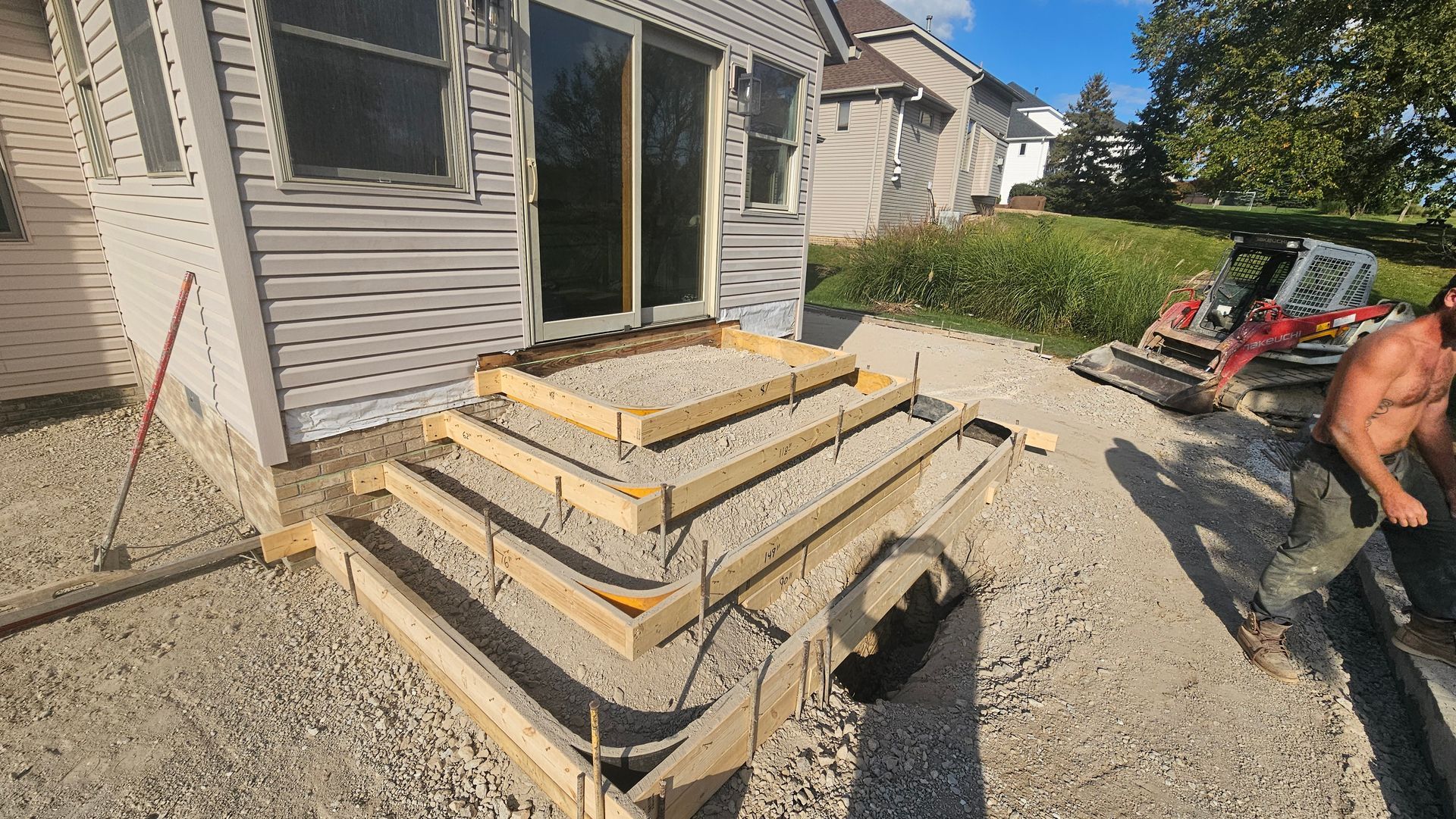 Wooden forms for concrete steps are built on a gravel base in front of a house, with a person working nearby.