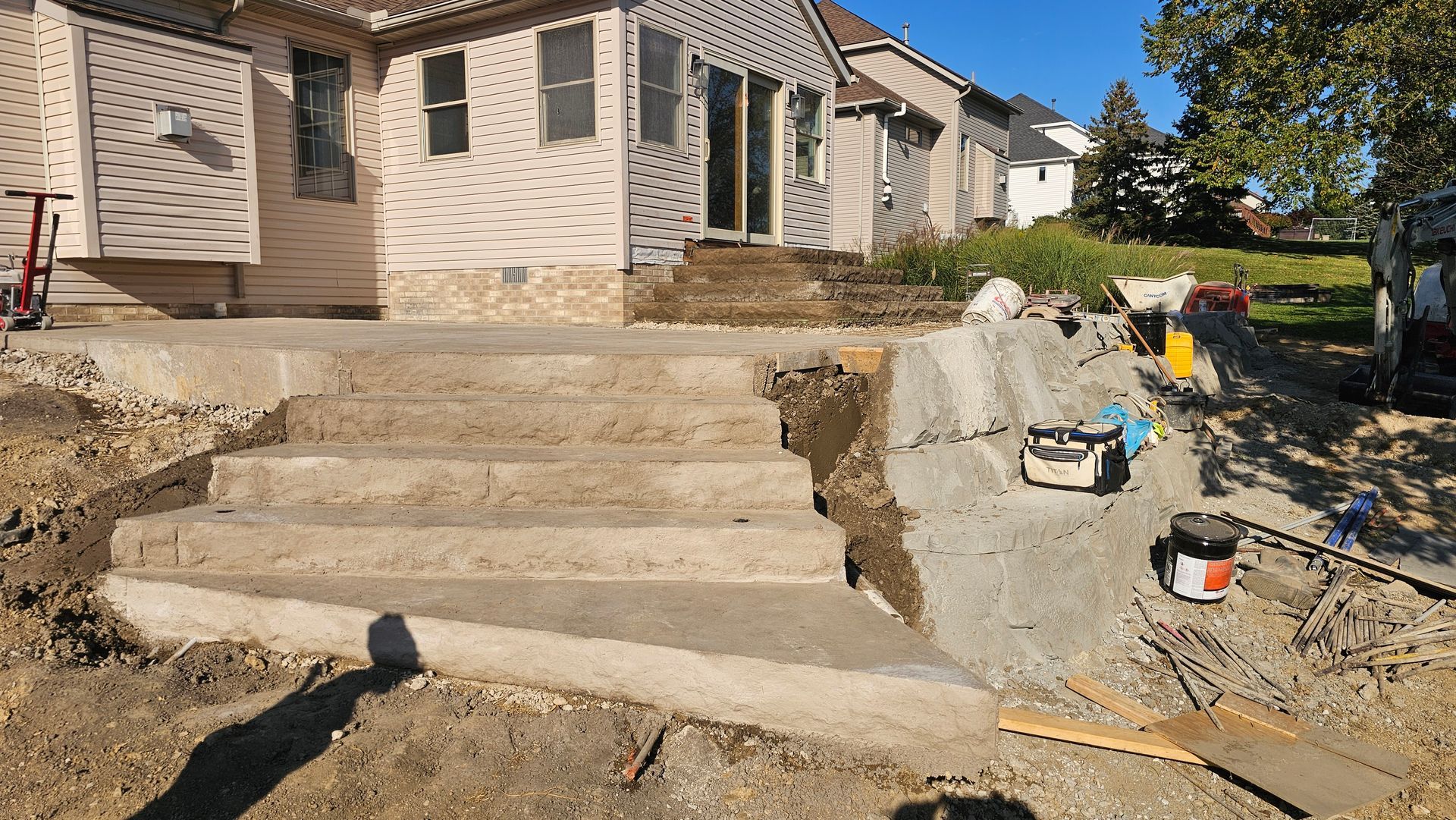 A residential backyard construction site featuring newly poured concrete stairs leading to the back of a beige house.