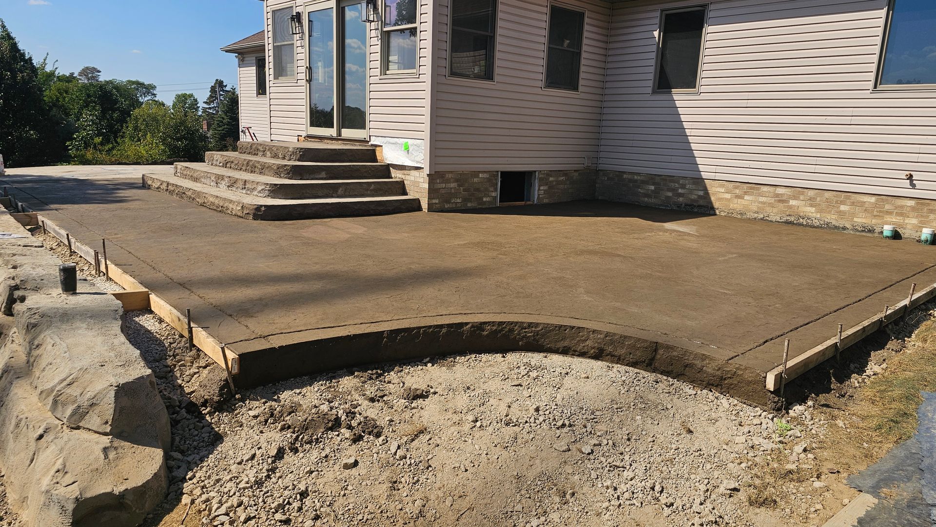 A concrete patio under construction in front of a house, featuring curved wooden forms and a gravel base.