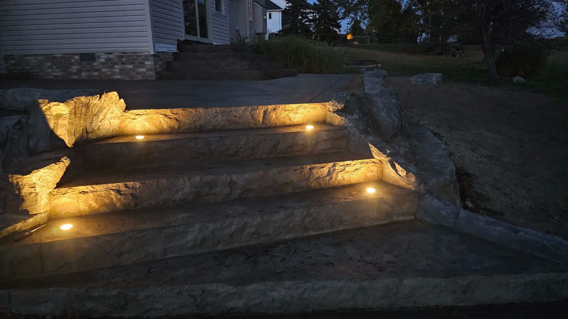 A stone staircase at dusk, illuminated by warm, round lights embedded in each step leading toward a house.