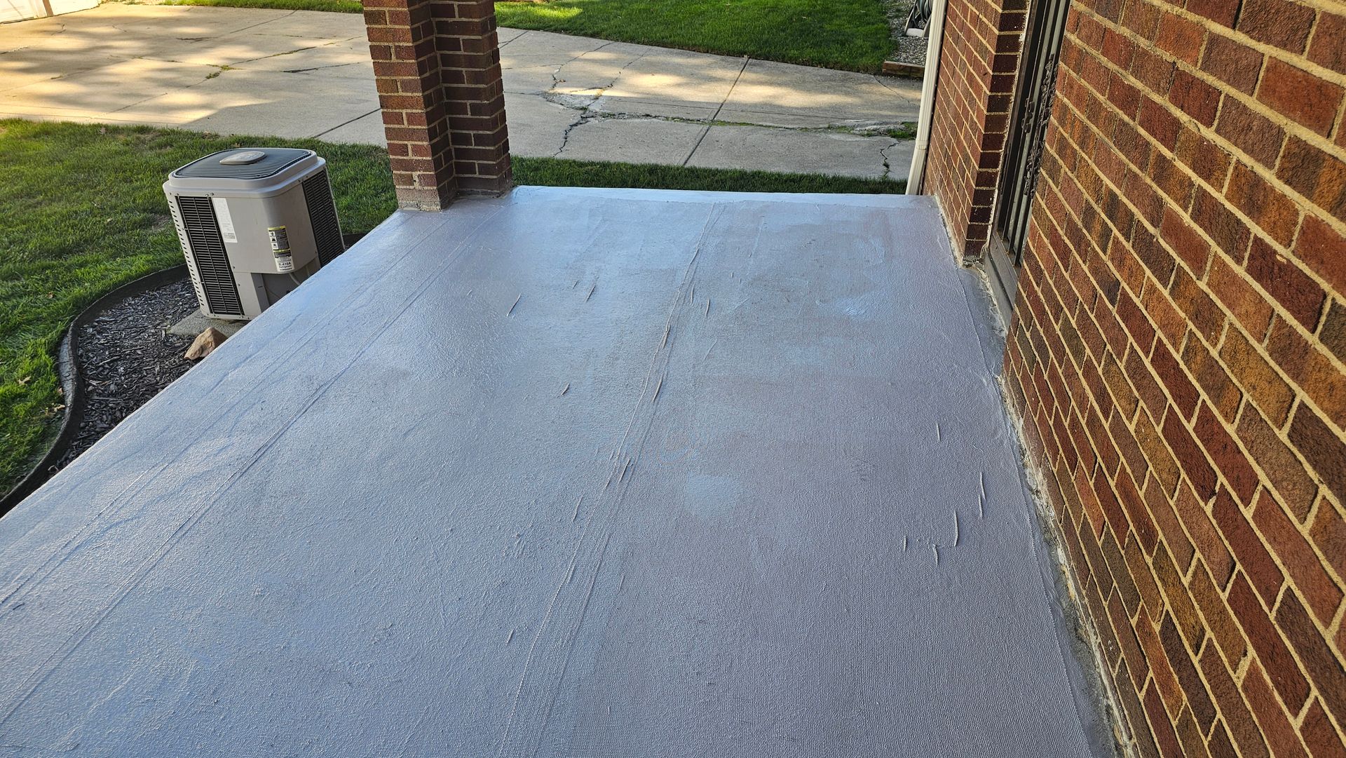 A textured gray porch surface viewed from an elevated angle, adjacent to a brick wall and a brick support pillar.