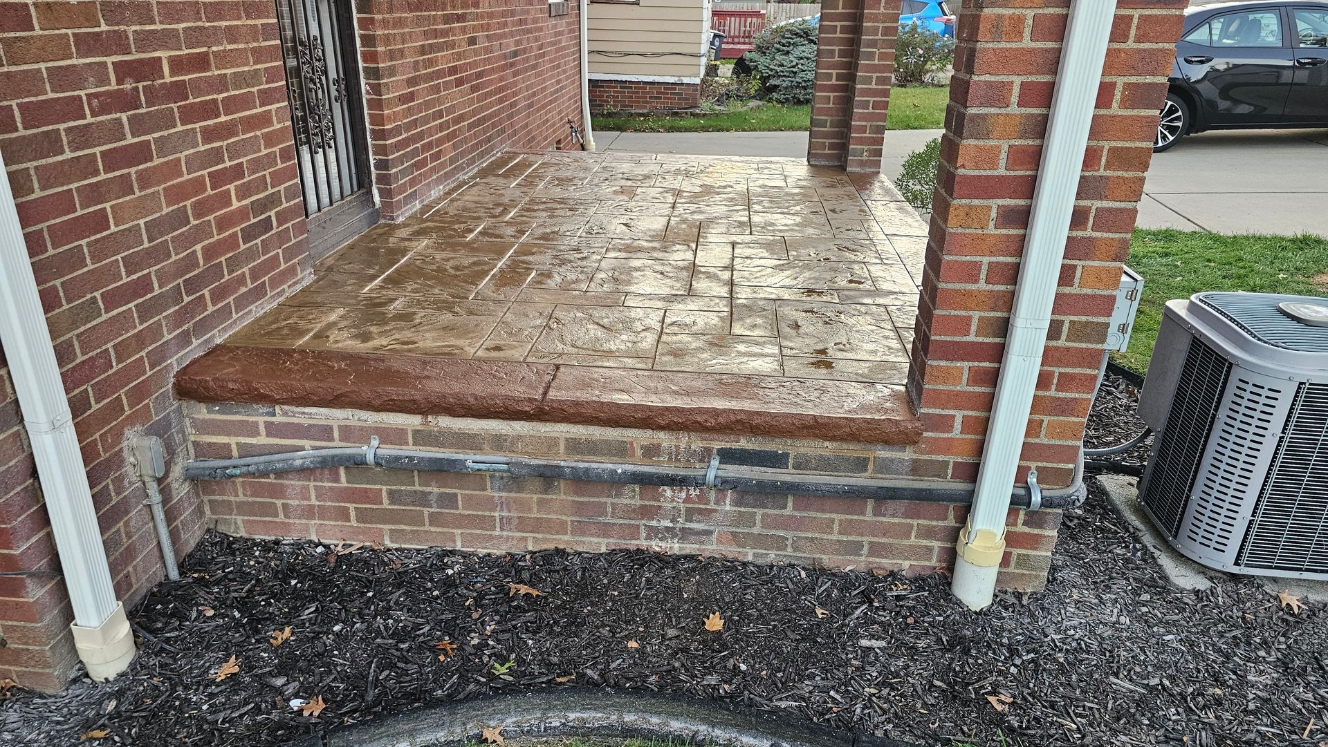 A stamped concrete porch with a brown brick facade and support columns, viewed from a landscaped front yard.