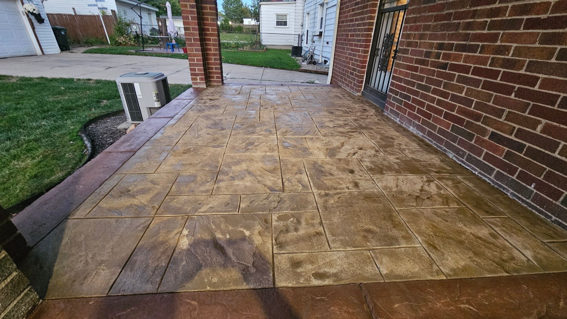 A brown, stamped concrete porch with a stone-like pattern, bordered by a dark frame next to a red brick house wall.