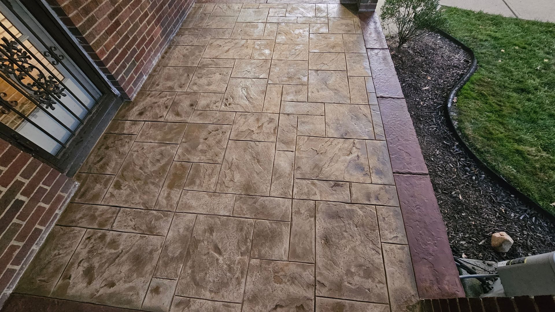 Tan stamped concrete porch with a rectangular pattern, bordering a garden bed with dark mulch and grass.