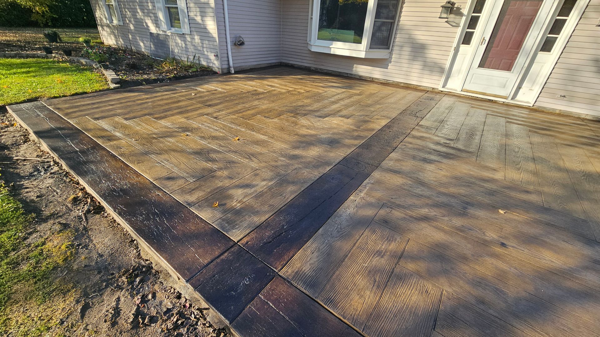 A new stamped concrete patio with a dark border, adjacent to a house with beige siding and a front entryway.