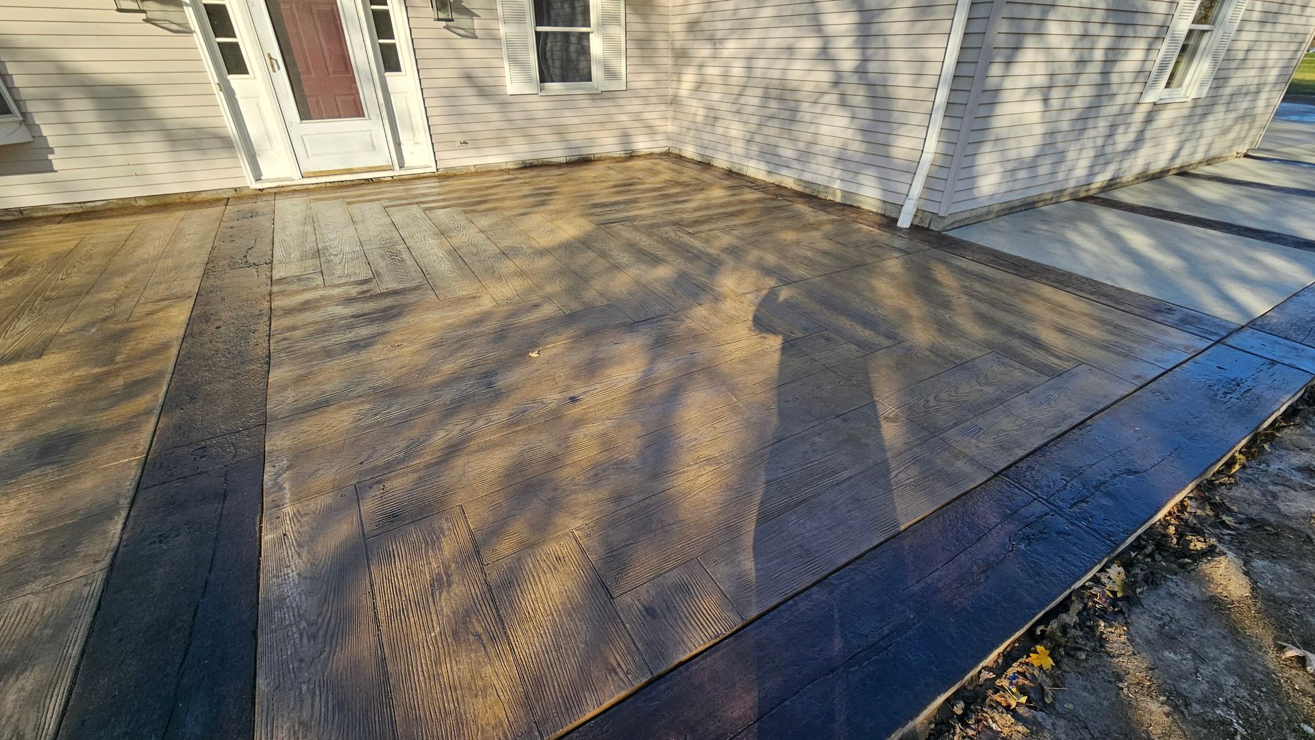 A freshly poured, textured concrete patio with a dark stained border extends from the side of a light-sided house.