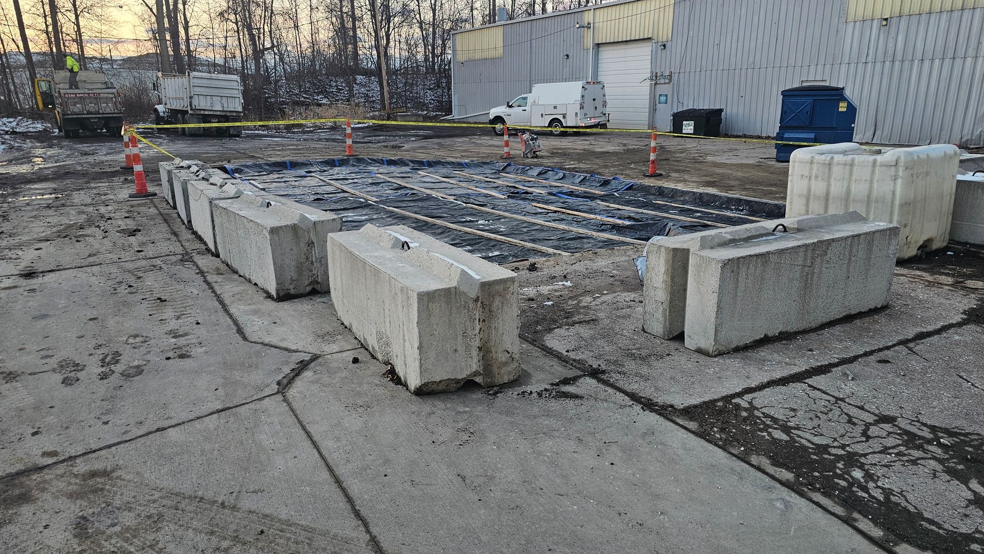 A construction site enclosed by concrete barriers and caution tape, featuring a dark ground patch and a white utility van.