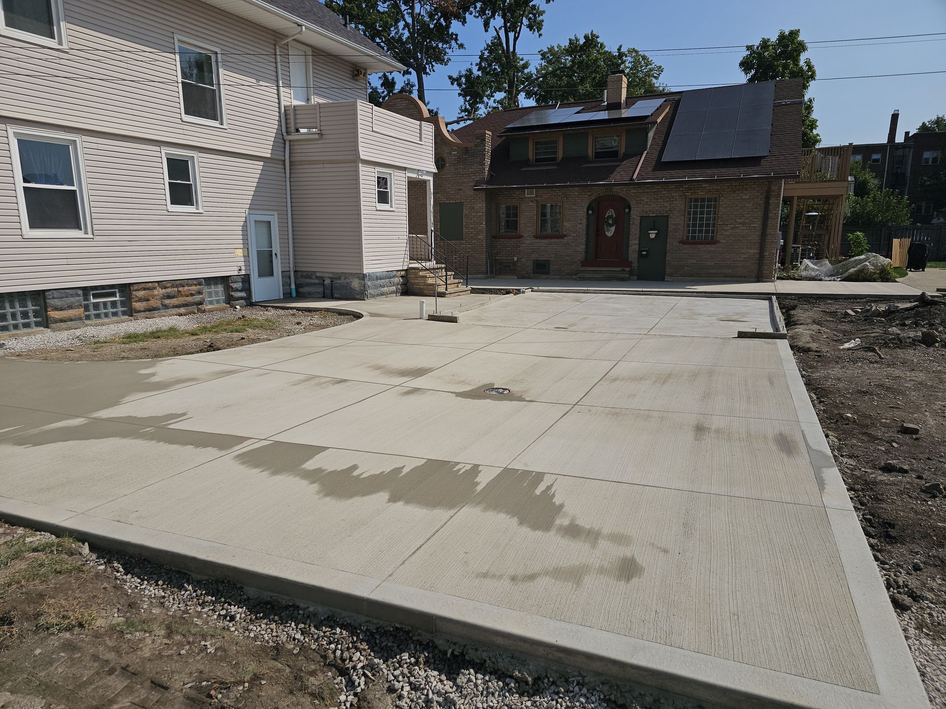 A newly poured concrete driveway sits between a light-sided house and a brick home on a sunny day.