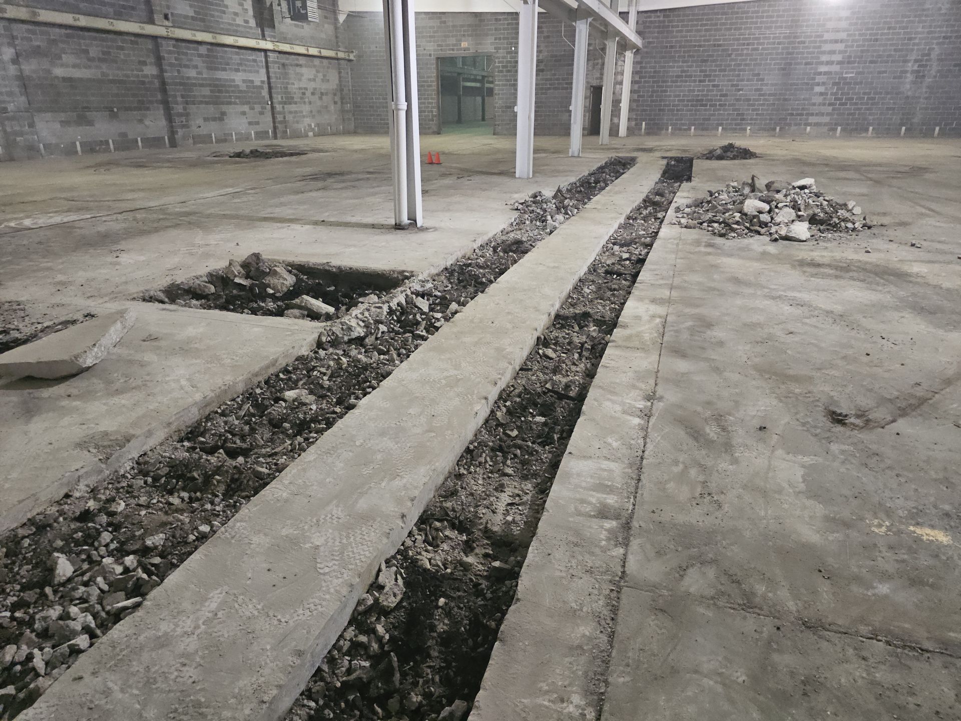 Concrete floor under construction featuring a long, narrow excavated trench with debris between support columns.