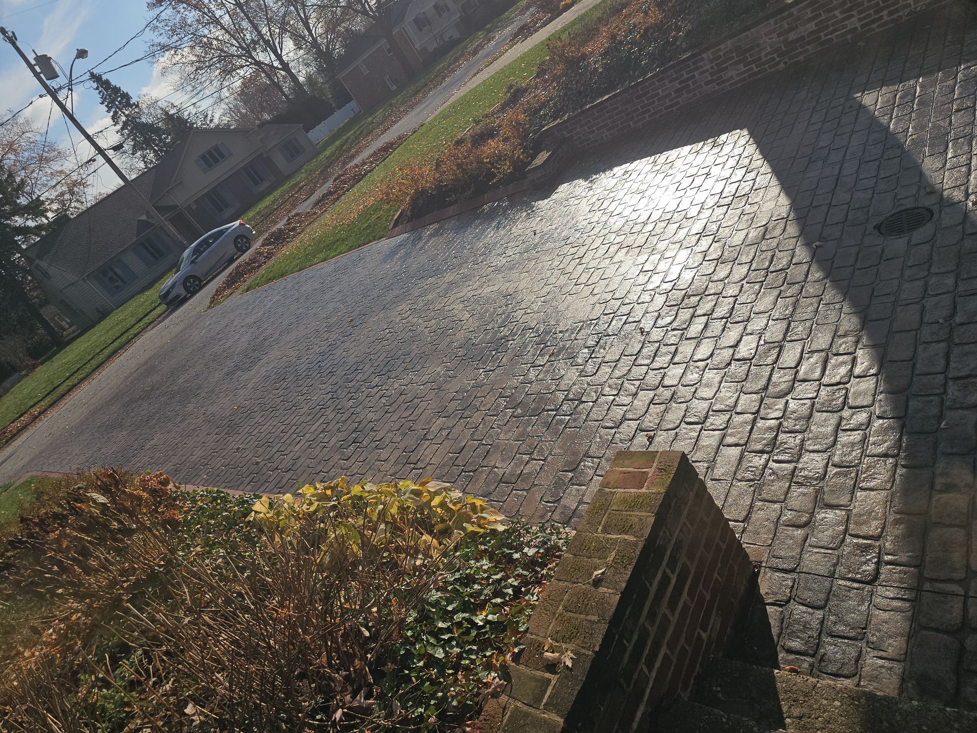 A wide, slightly angled view of a driveway paved with grey cobblestones, flanked by a brick wall and autumn bushes.