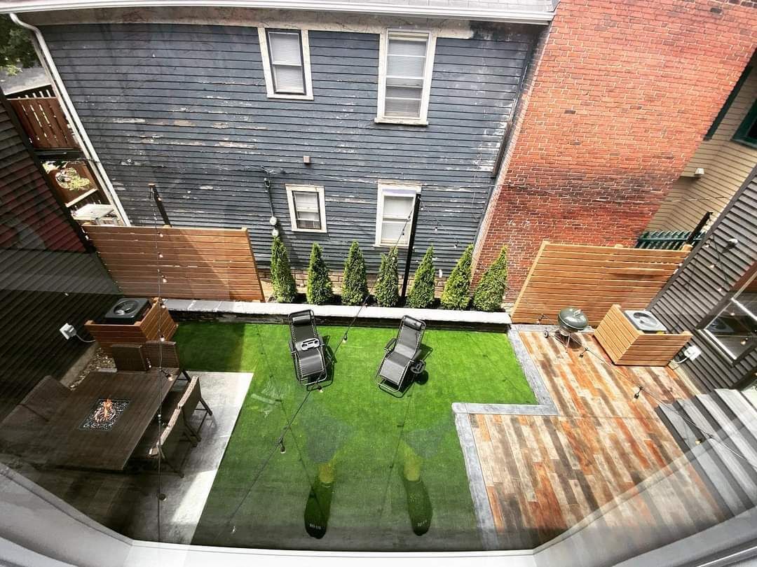 An urban backyard with artificial turf, two lounge chairs, a wooden dining set, a brick wall, and small evergreen trees.