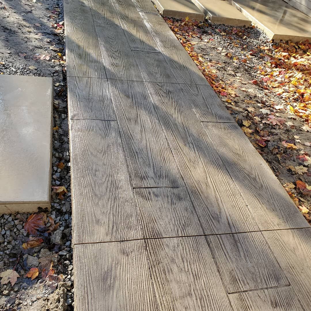 Textured, gray concrete pavers resembling weathered wooden planks laid out on a gravel and leaf-covered ground.