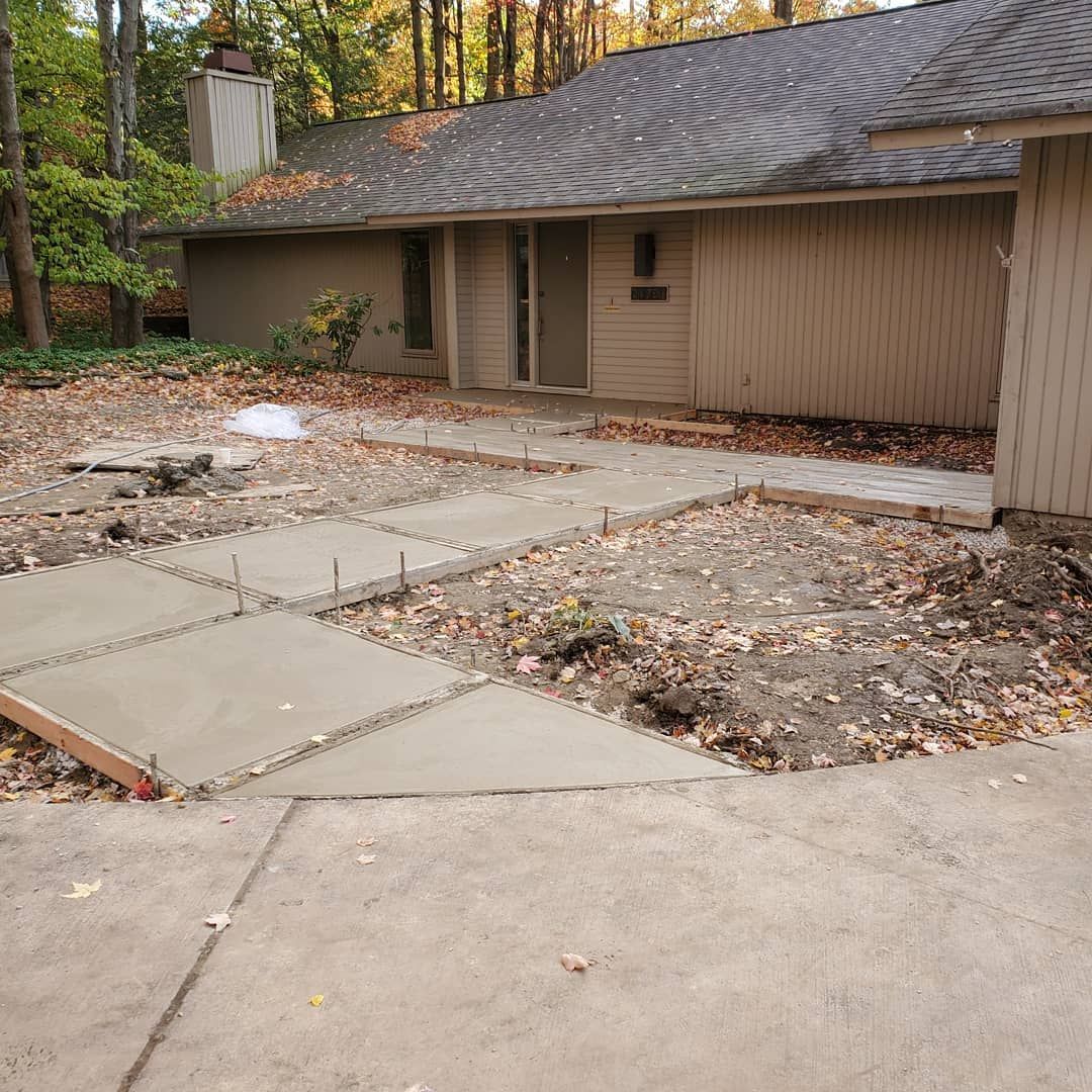 Freshly poured concrete walkway slabs in a residential yard leading toward a house entrance with wood-paneled siding.