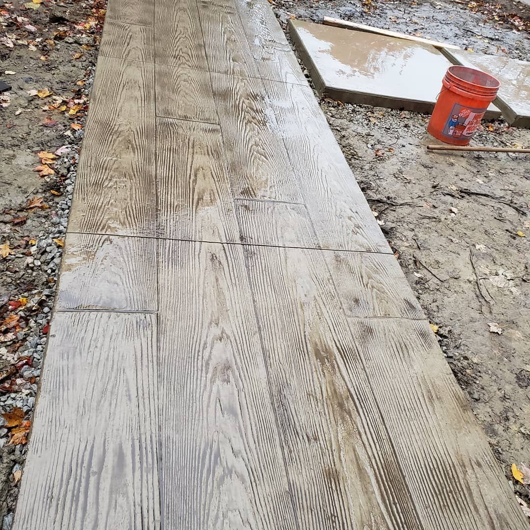Newly poured stamped concrete walkway with a wood grain texture, next to an orange bucket and a rectangular form.