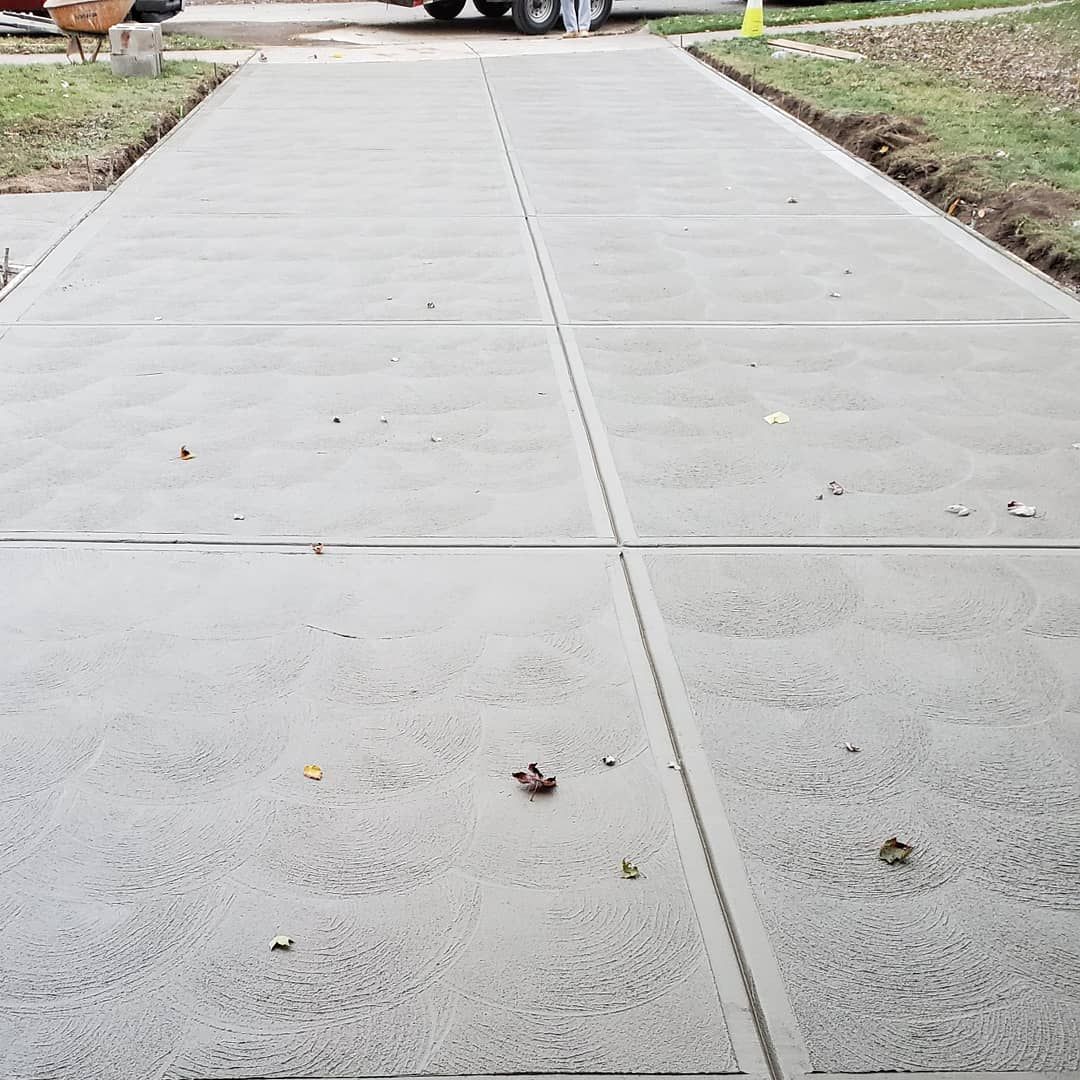 A newly poured concrete driveway featuring a uniform grid pattern of square slabs, extending toward a vehicle in the rear.