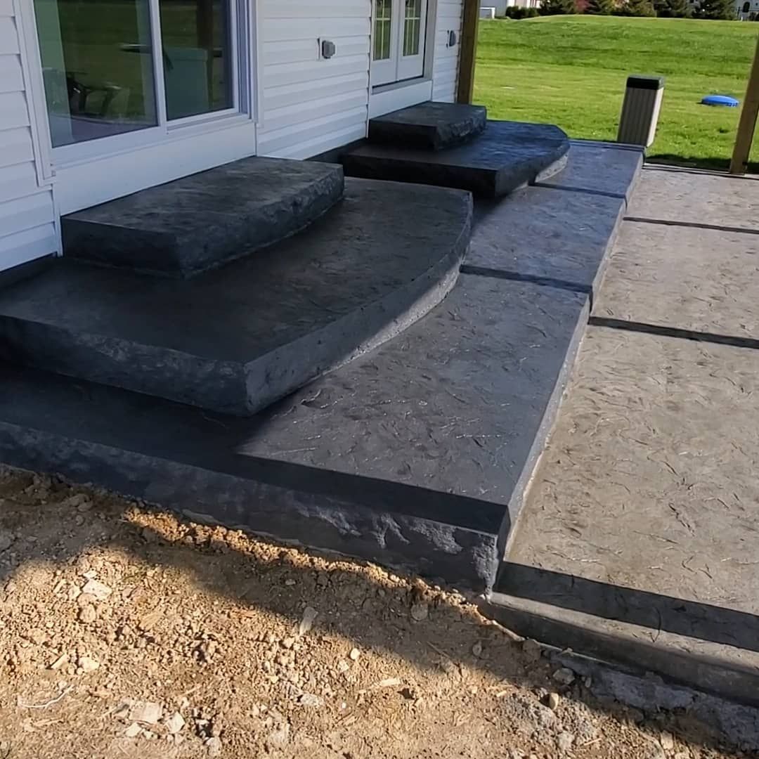 Dark grey stamped concrete steps leading up to two building entrances, beside a patio on a dirt ground surface.