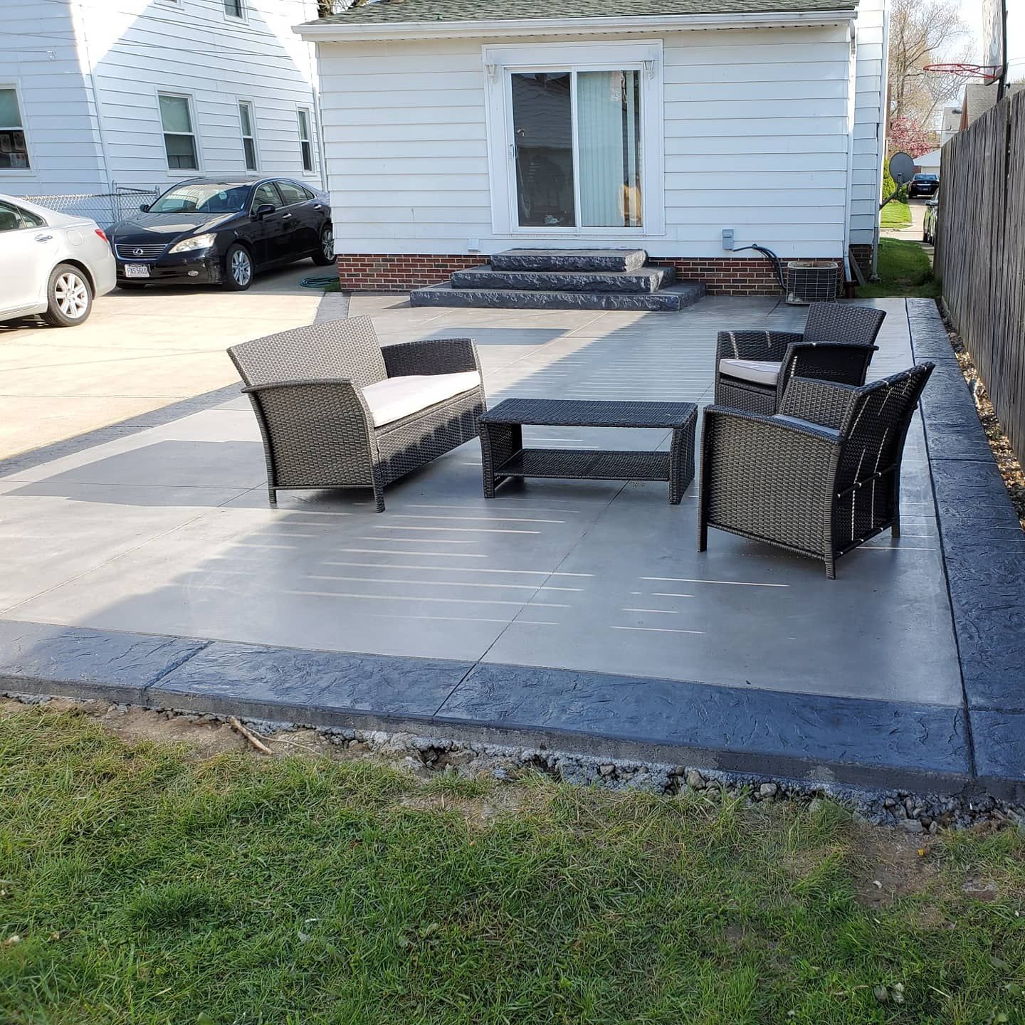 A gray concrete patio with dark stone borders featuring wicker patio furniture outside a white house with sliding doors.