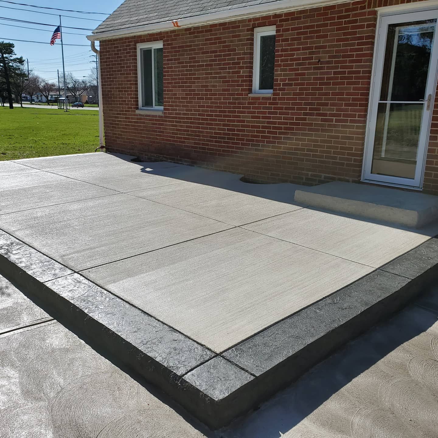 A newly poured concrete patio with a dark grey textured border adjacent to a brick house and a white door.
