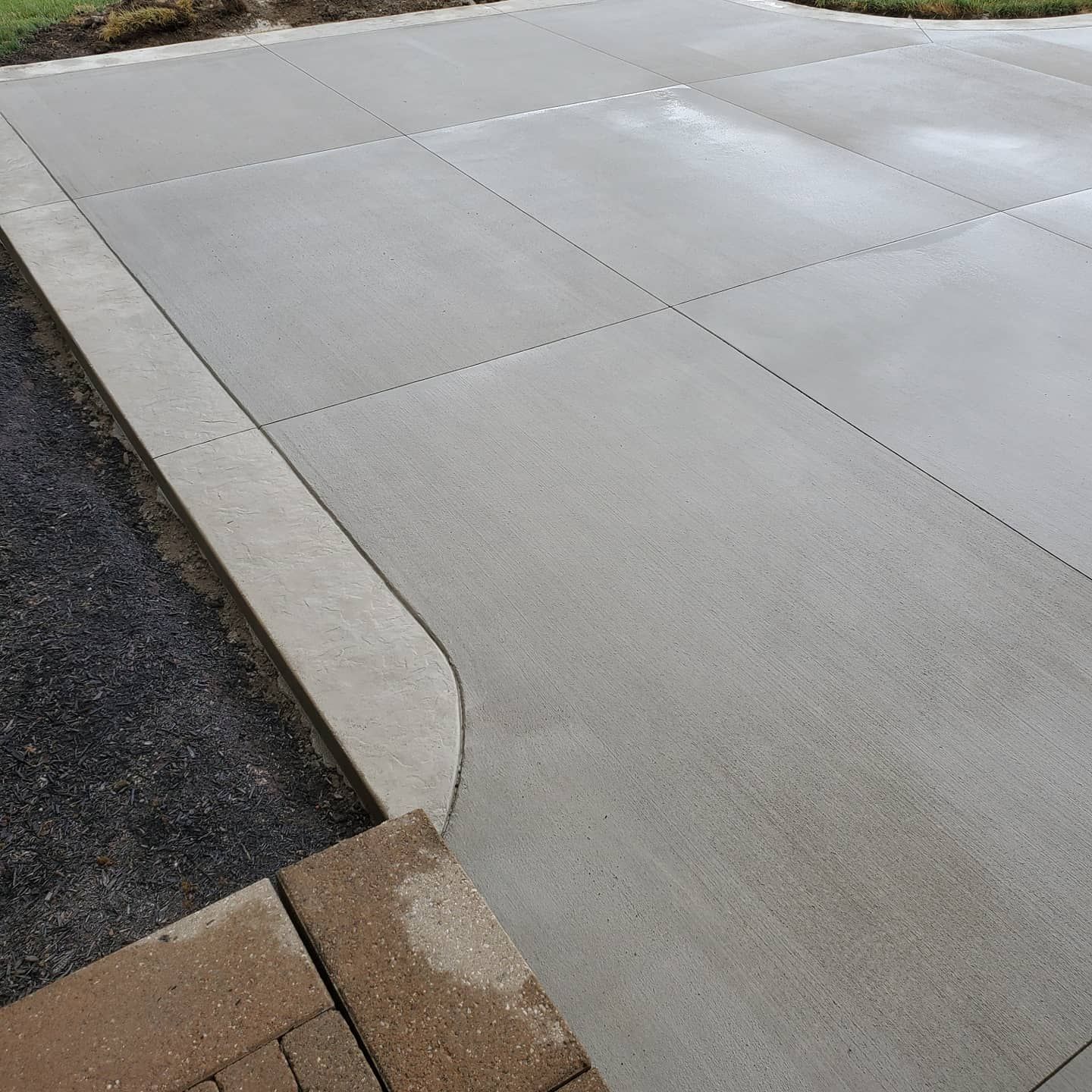 A new concrete driveway with a curved edge, adjacent to a section of brown stone pavers and a dark gravel border.