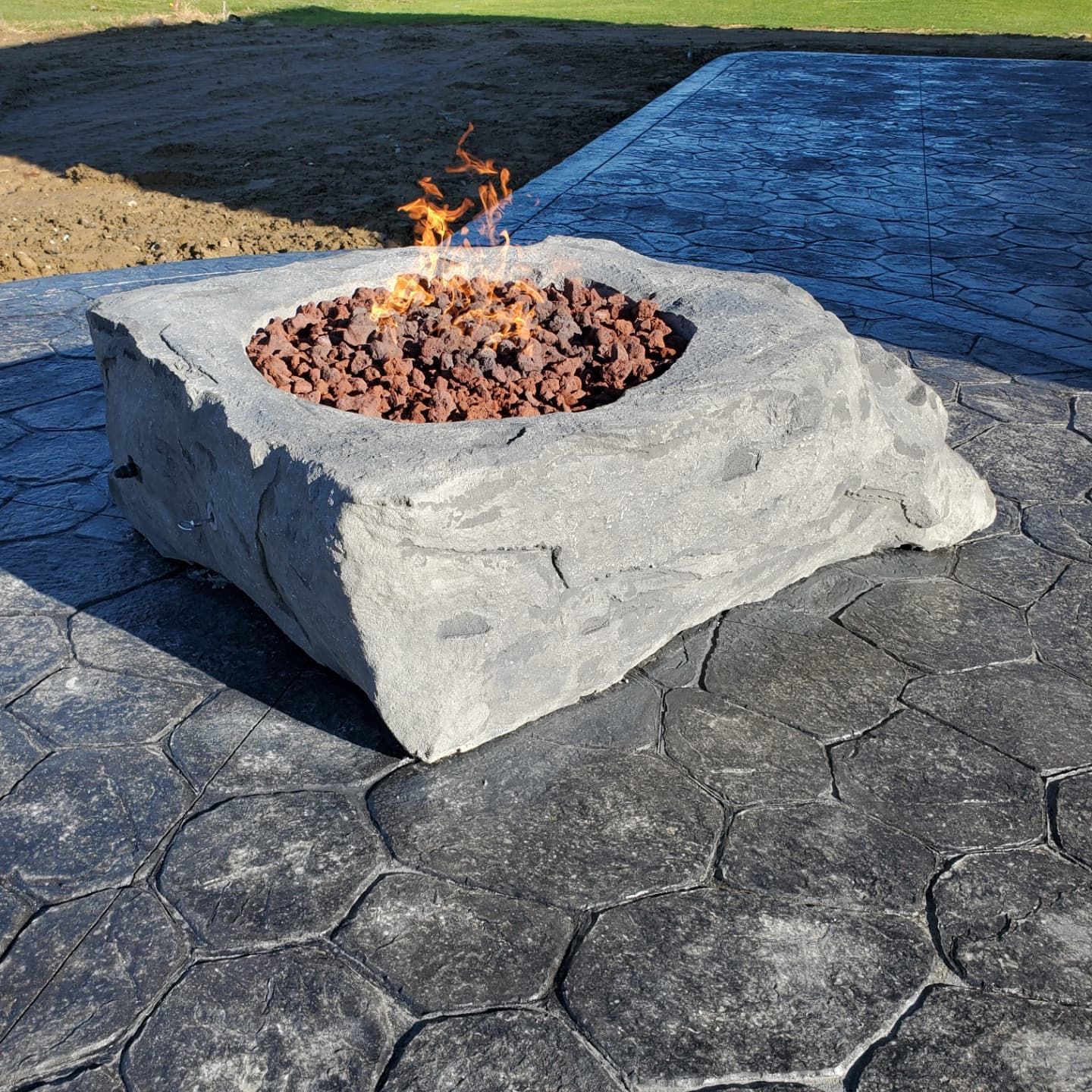 A square, stone-textured outdoor fire pit with flickering flames sits on a dark, patterned concrete patio.