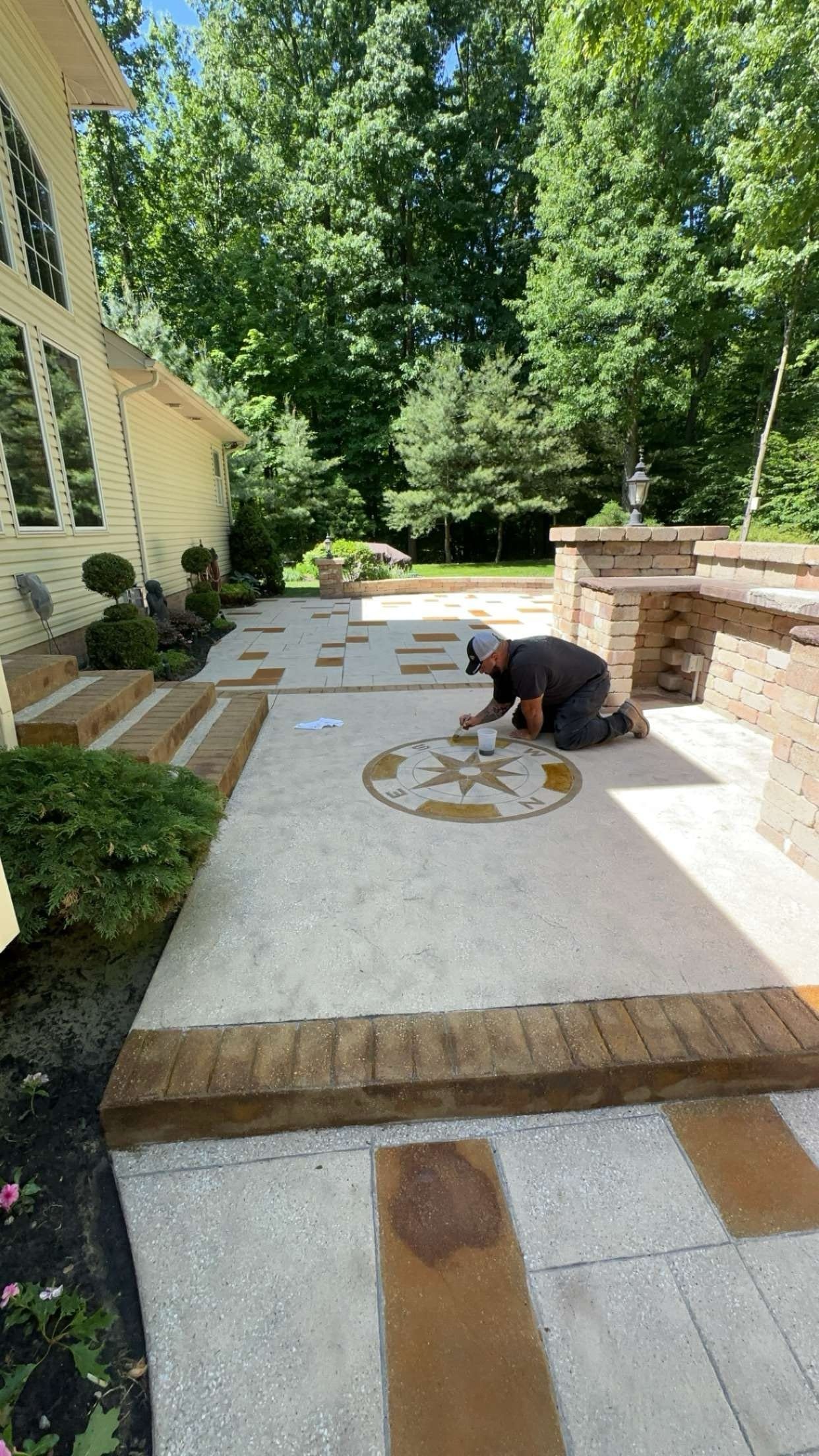 A contractor installs a stone compass inlay into a light-colored paver patio next to a house exterior.