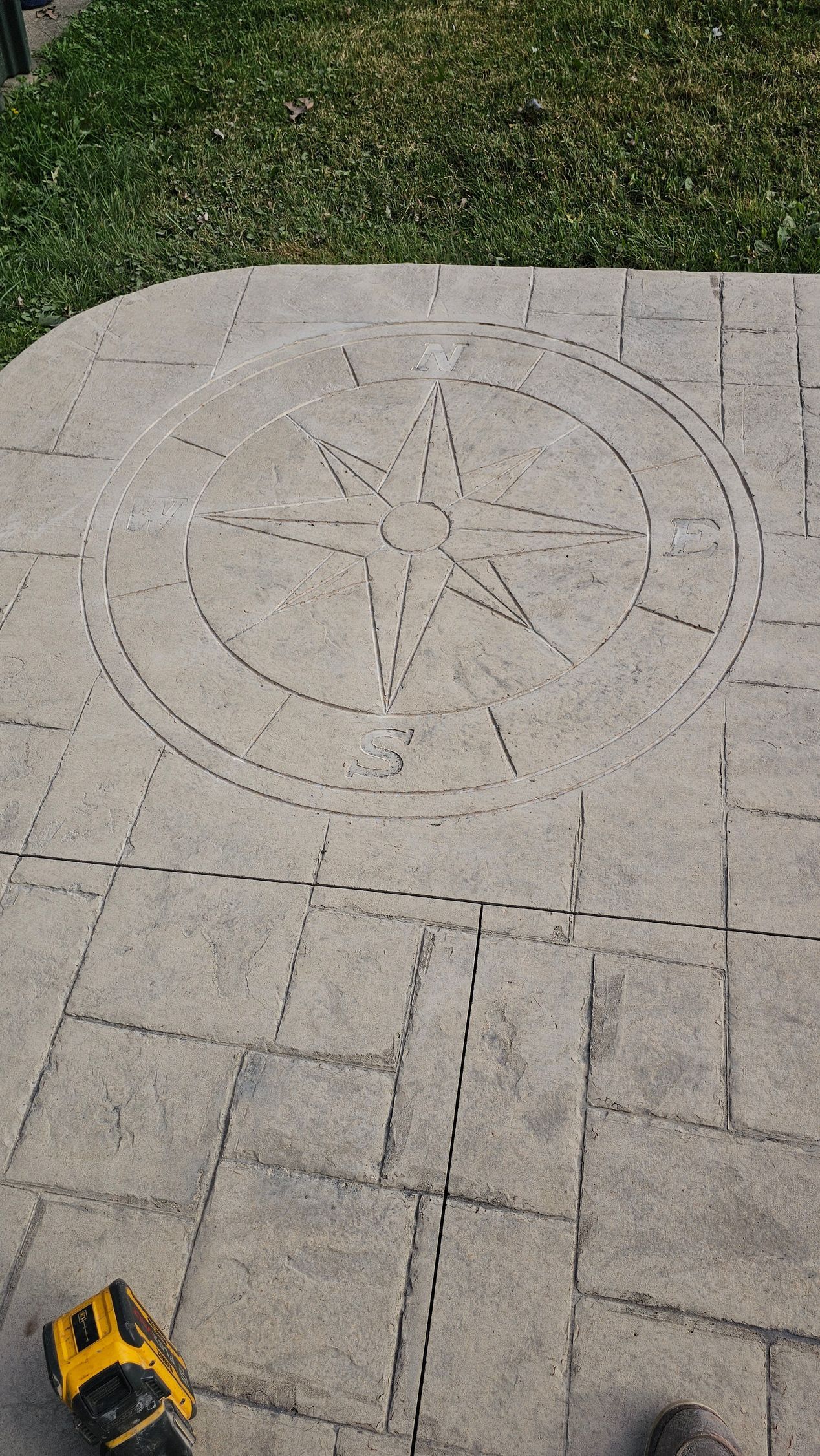 A compass rose design marked with dotted lines on light gray patio pavers, with a small yellow power tool nearby.