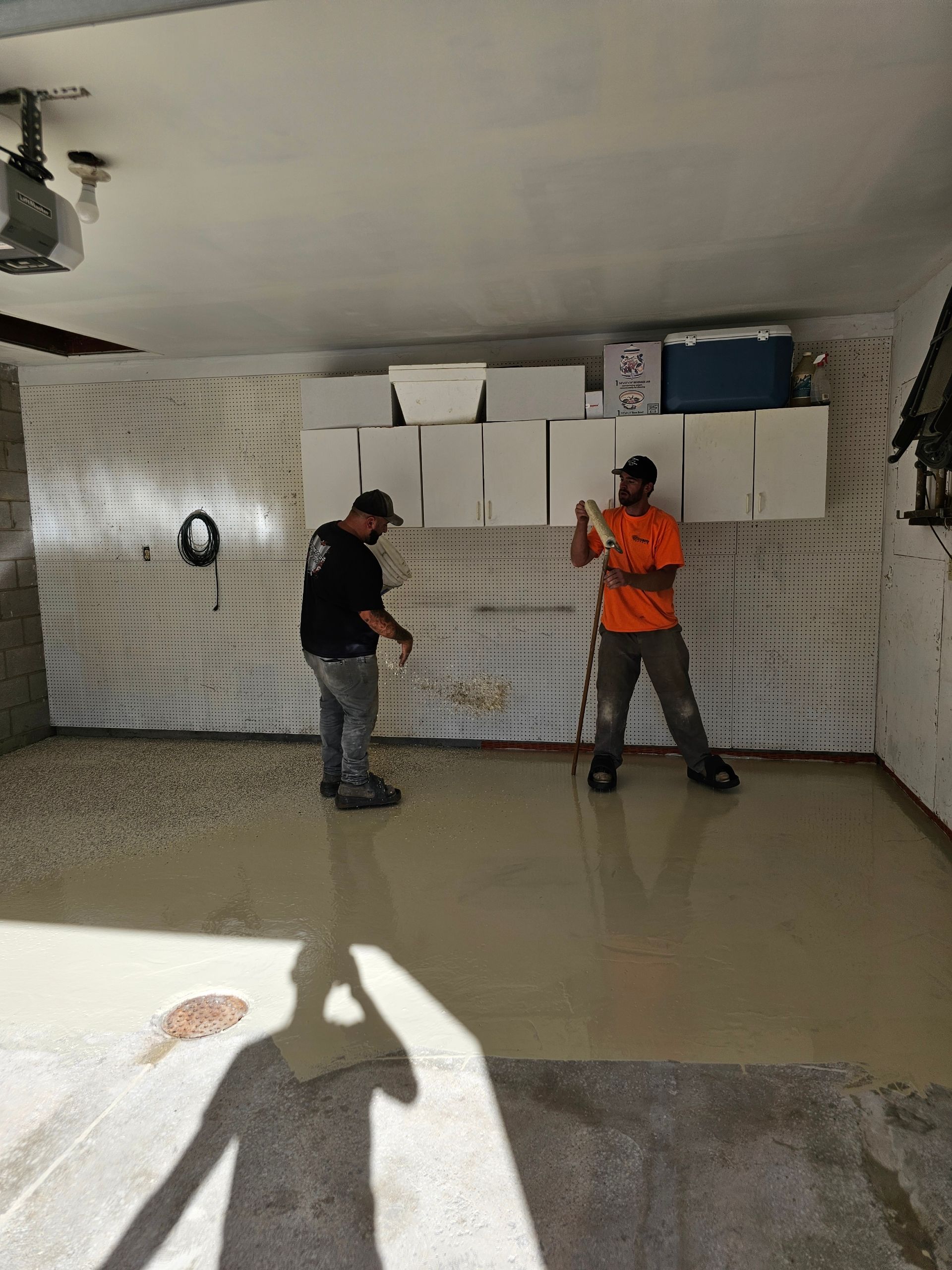 Two people in a garage with white wall panels work on a freshly poured concrete floor.