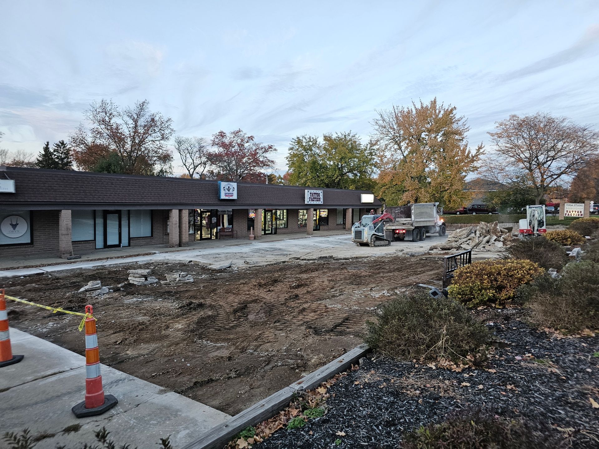 Construction equipment sits on an excavated dirt lot in front of a small commercial strip mall under an autumn sky.