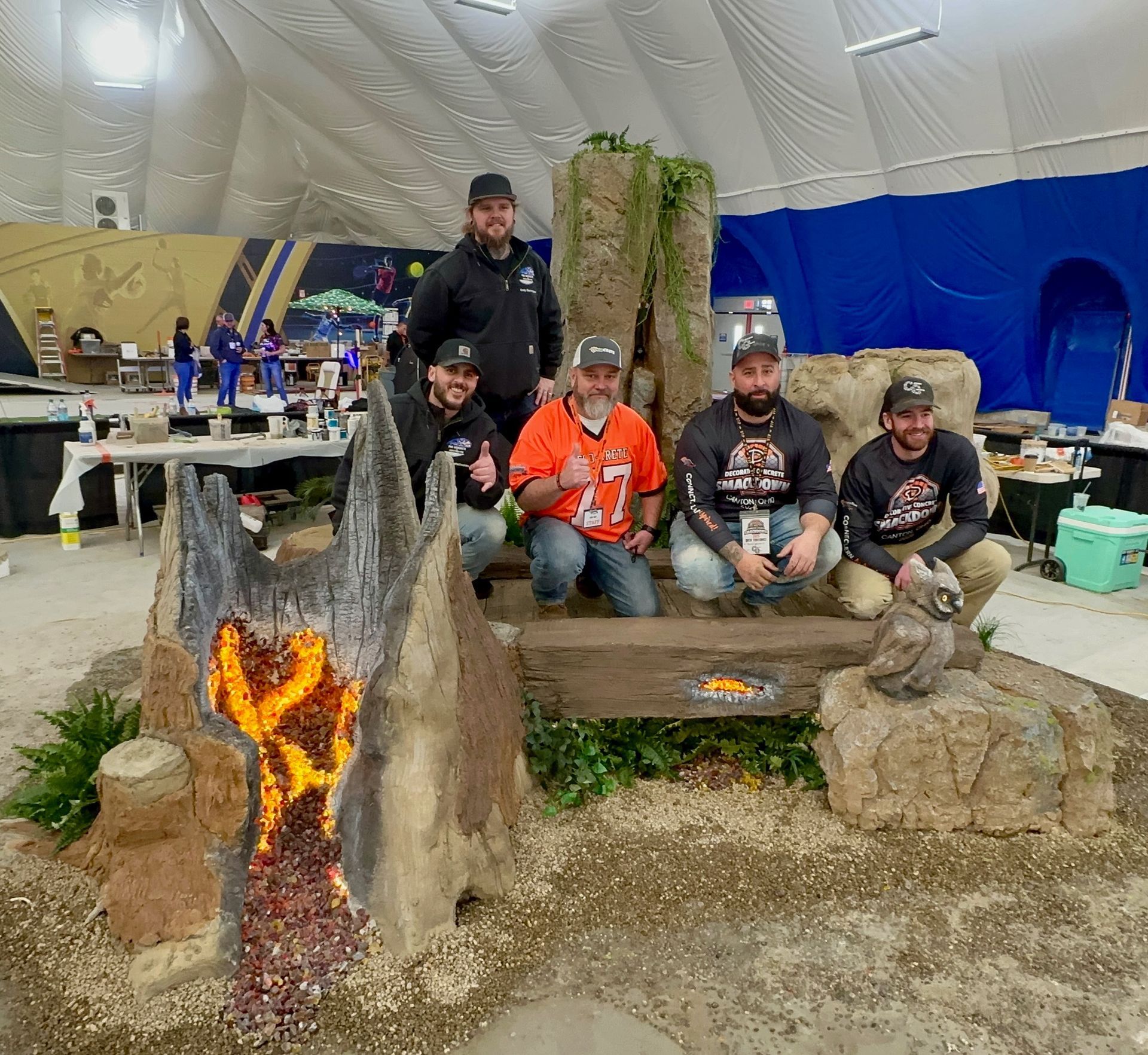 Five people pose behind a large, illuminated artificial volcano and rock display inside an event tent.