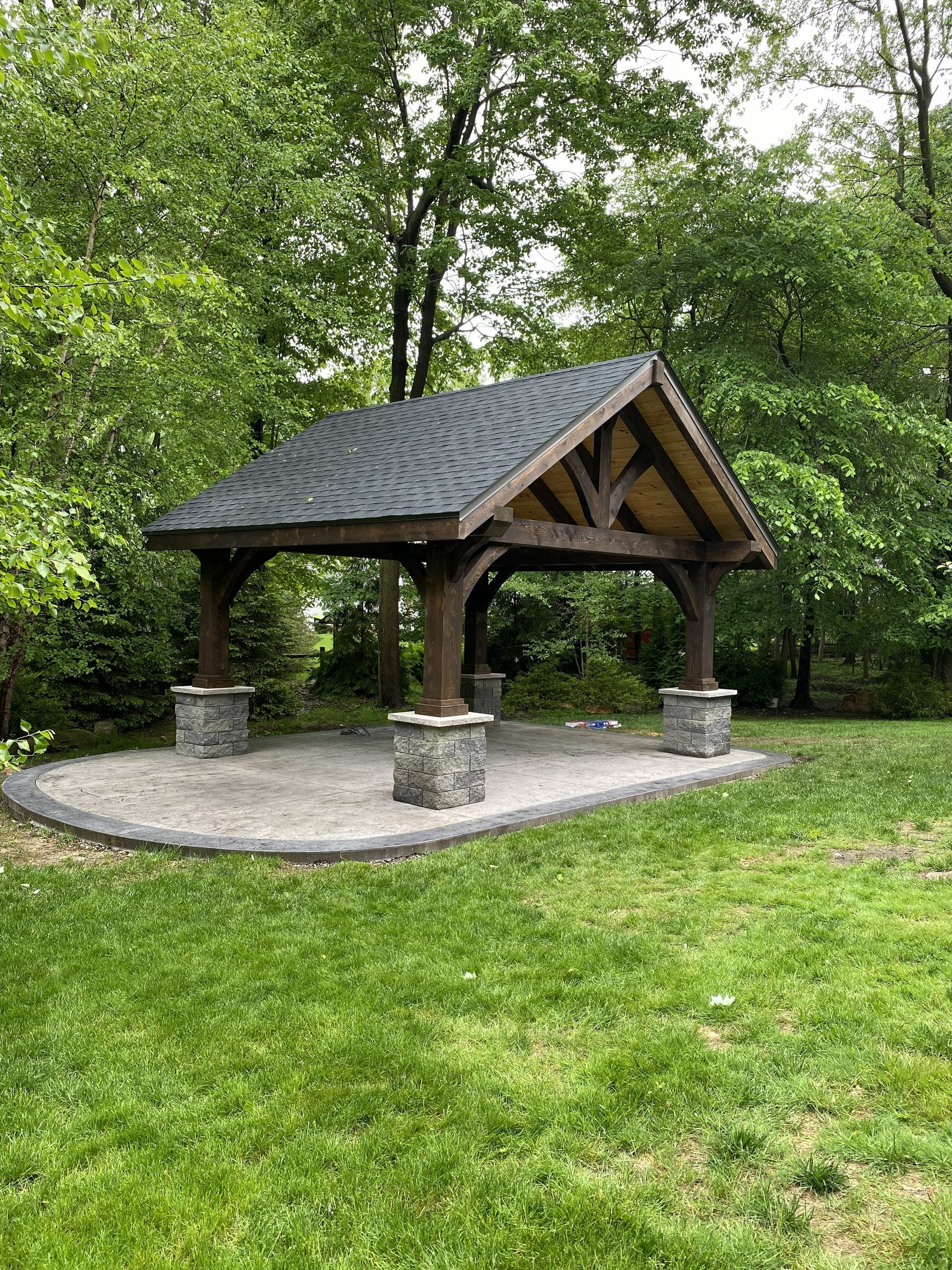 A dark wood timber-frame gazebo with a shingled roof and stone pillar bases, set on a gravel pad in a grassy yard.