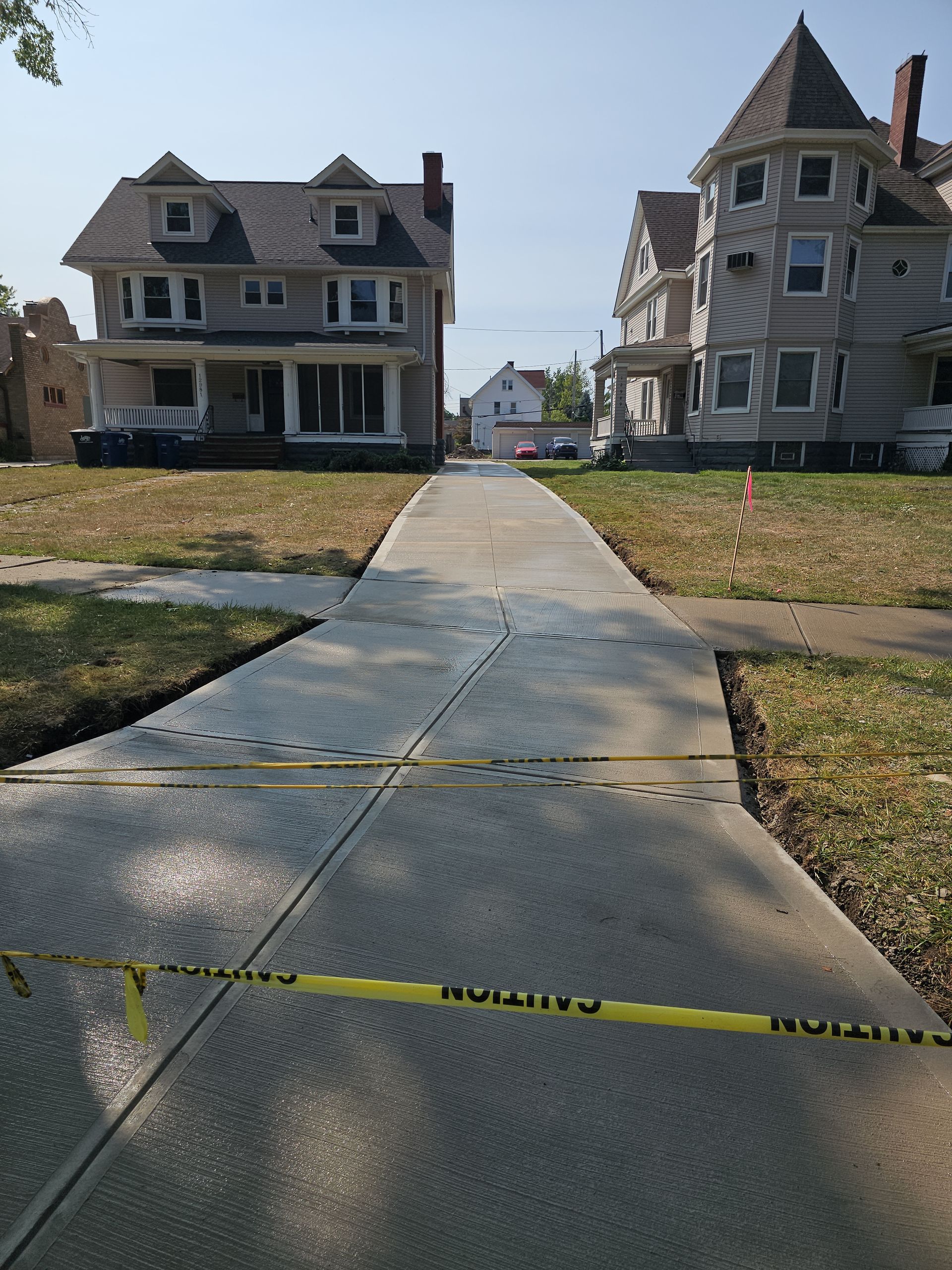 A newly poured concrete driveway leads between two houses, blocked off by yellow caution tape.